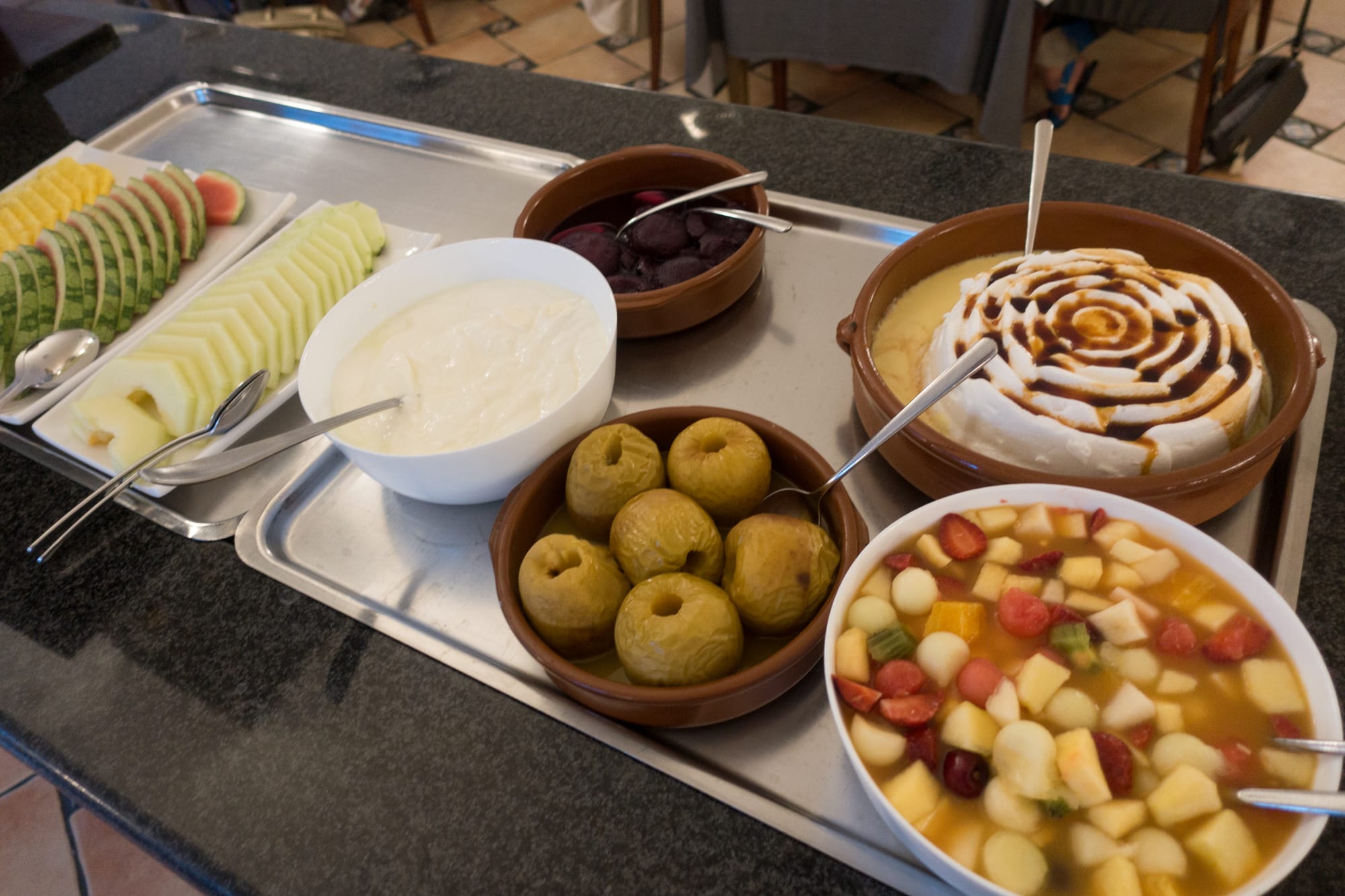 A tray of desserts including sliced melon and watermelon, a bowl of plain white cream, a dish of stewed fruit, and a round dish of custard with a caramelised swirl pattern on top