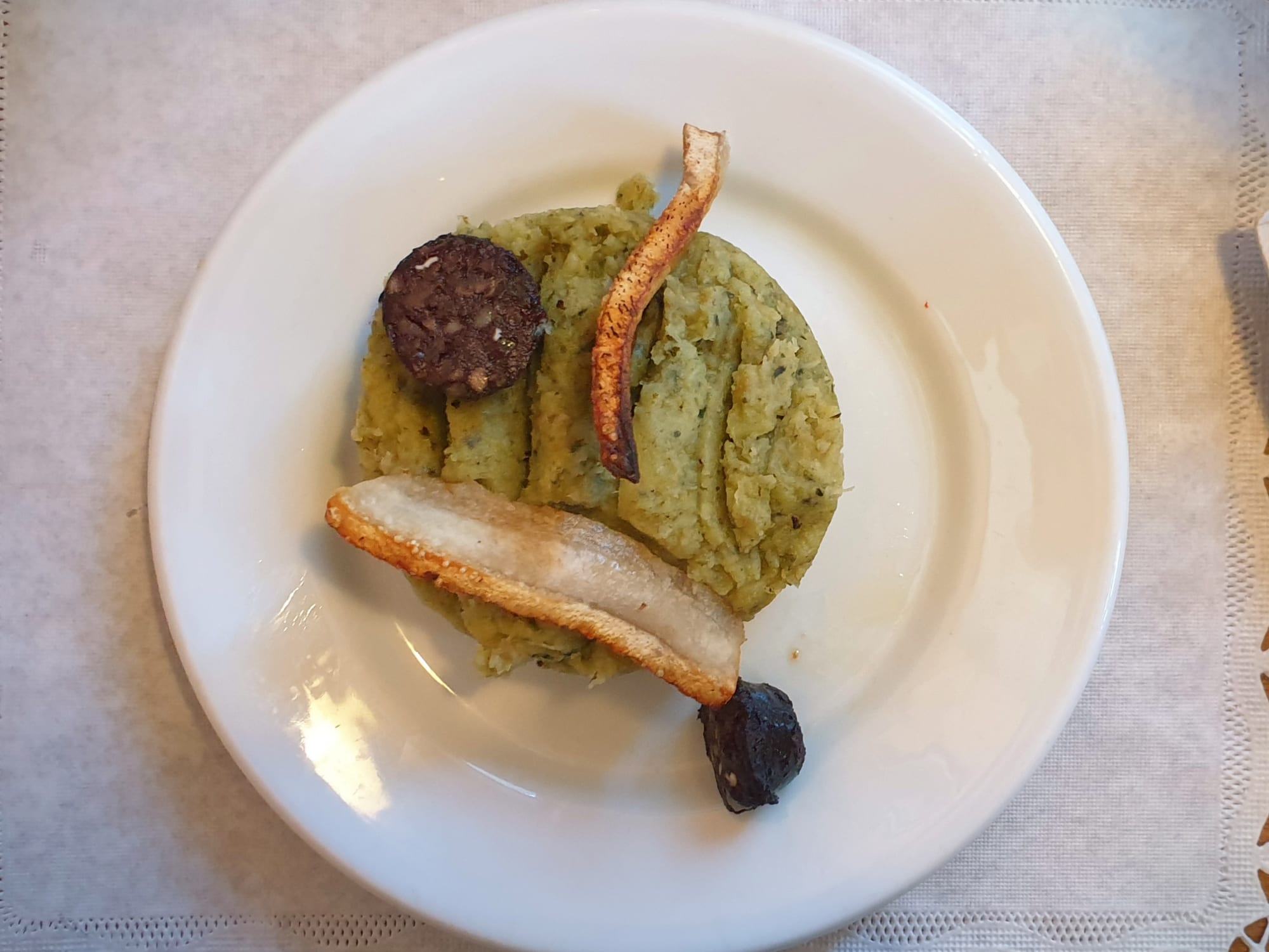 A plate of mashed green cabbage and potato with a round slice of dark blood sausage, a strip of crisped pork skin and a piece of roasted garlic