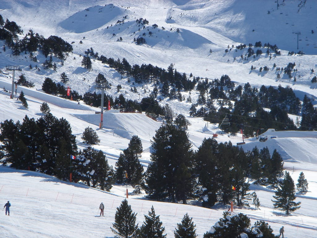 Skiers on a wide groomed piste below a ridge of brown-grey Pyrenean peaks with patchy snow