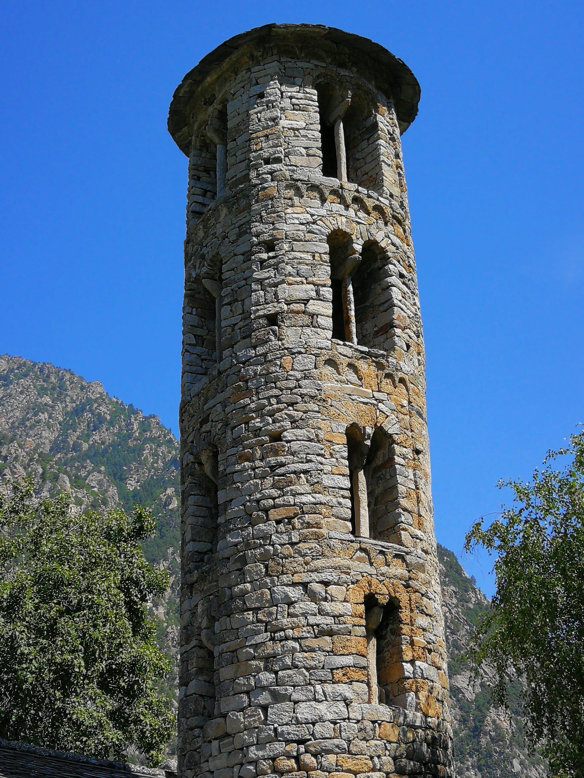 A small whitewashed stone church with a tall, round, four-storey bell tower, against a wooded hillside
