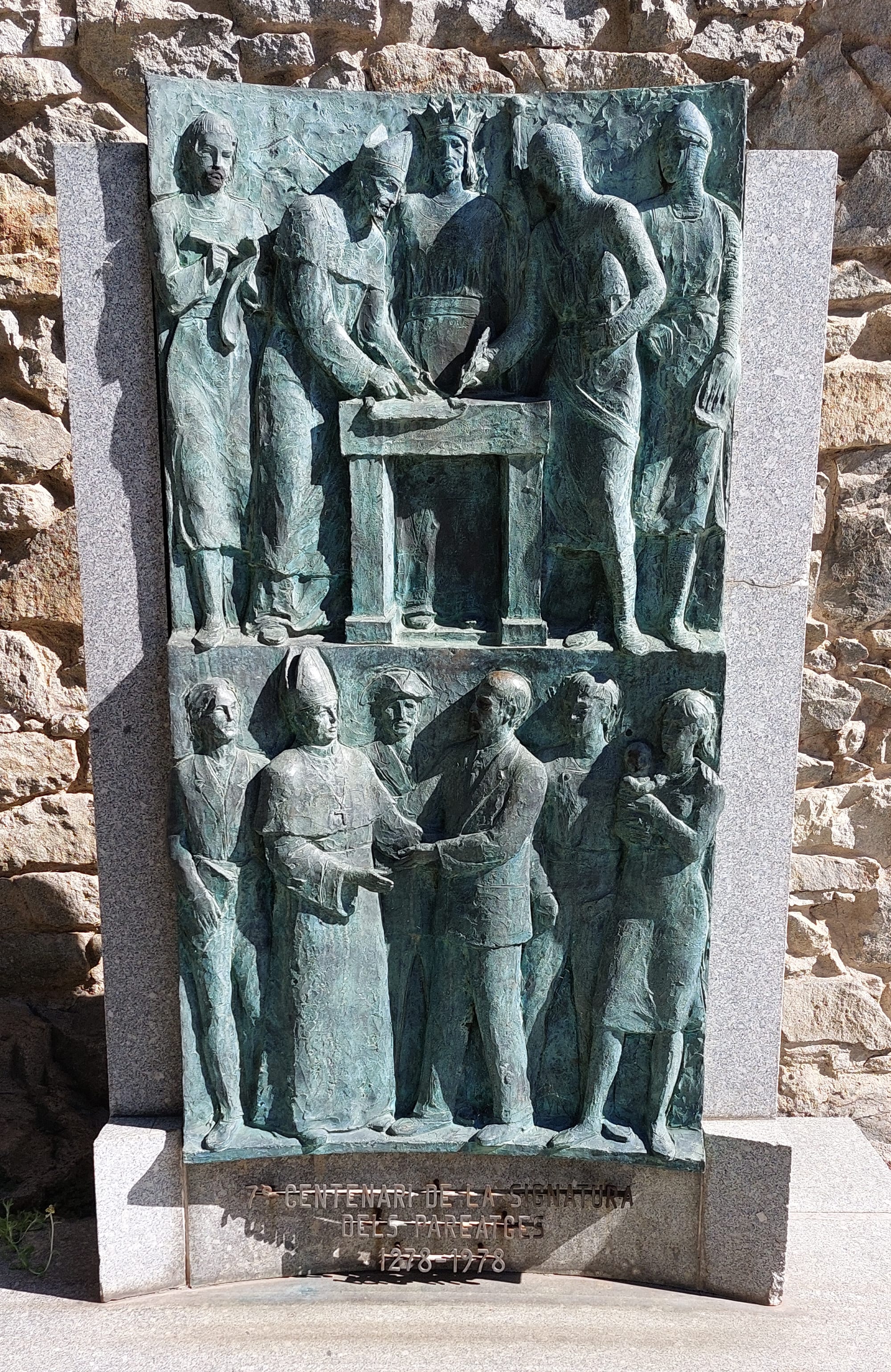 A green bronze relief in two panels set against a stone wall: above, a bishop and a knight in chain mail sign a document at a table; below, a bishop in a mitre shakes hands with a man in a suit