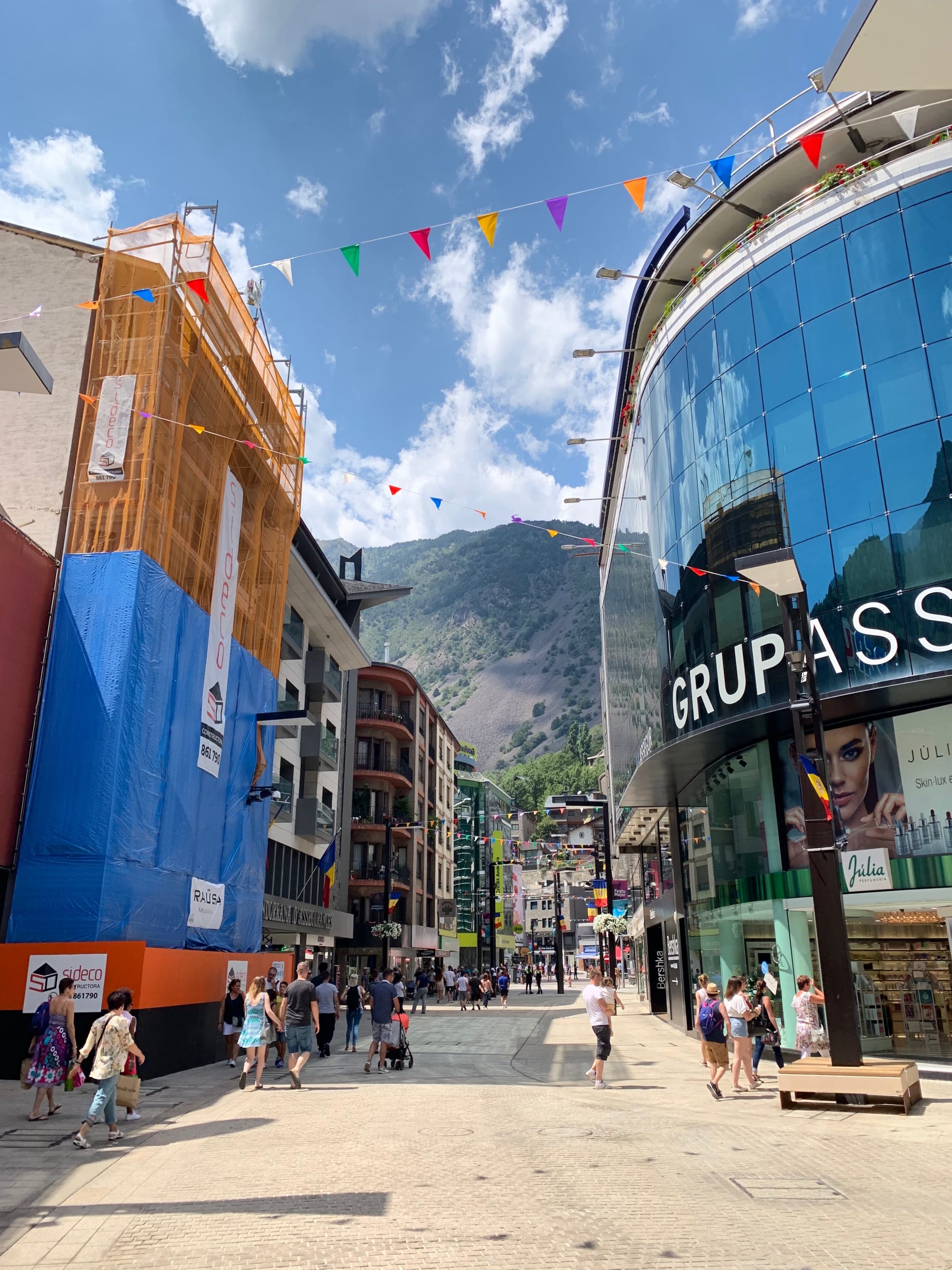 A busy pedestrian shopping street in Andorra la Vella lined with stores, with mountains rising behind the buildings