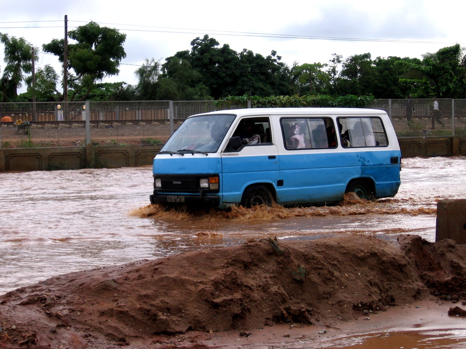 A blue-and-white candongueiro minibus with passengers crossing a flooded street in Luanda after heavy rain