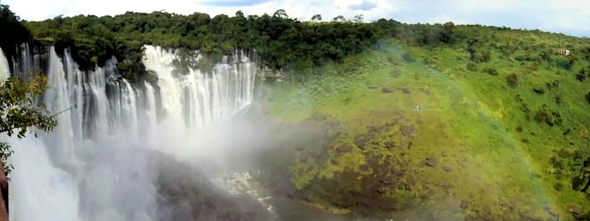 A very wide curving waterfall dropping over a forested escarpment into a misted gorge