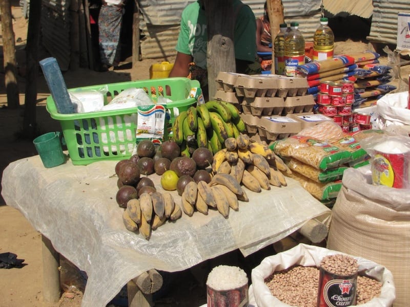 An outdoor market stall piled with plantains, avocados, bottles of cooking oil, eggs and sacks of dried beans and rice