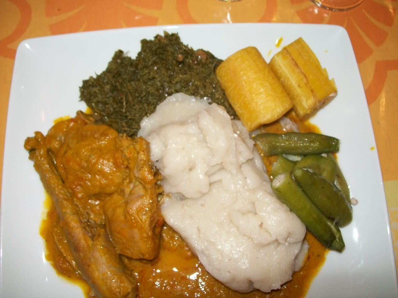 A square white plate with chicken in orange palm-oil sauce, a mound of white starch, dark green cooked leaves, sliced plantain and okra pods
