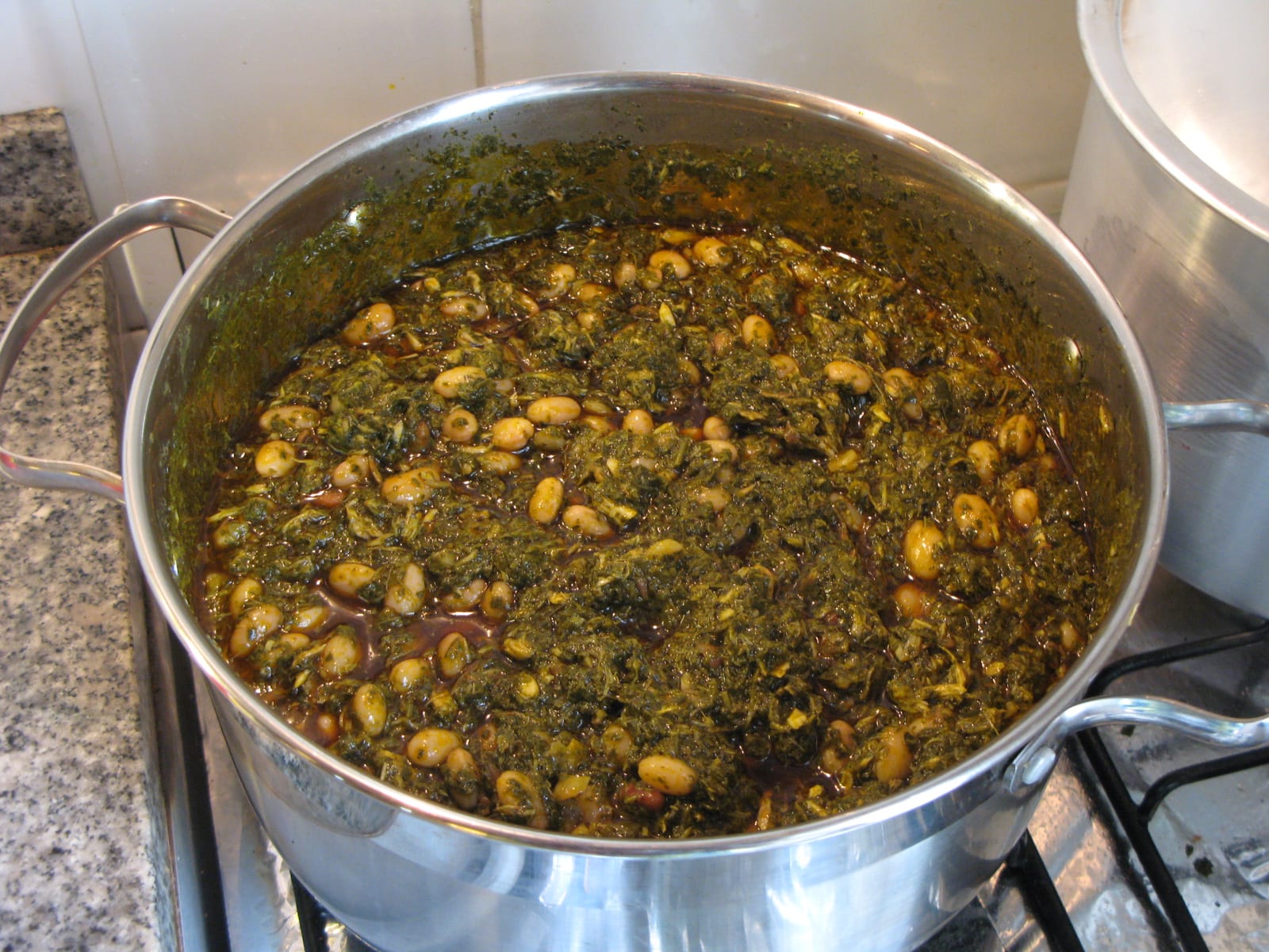 A large steel pot on a stove full of dark green cooked cassava leaves mixed with pale beans in an oily sauce
