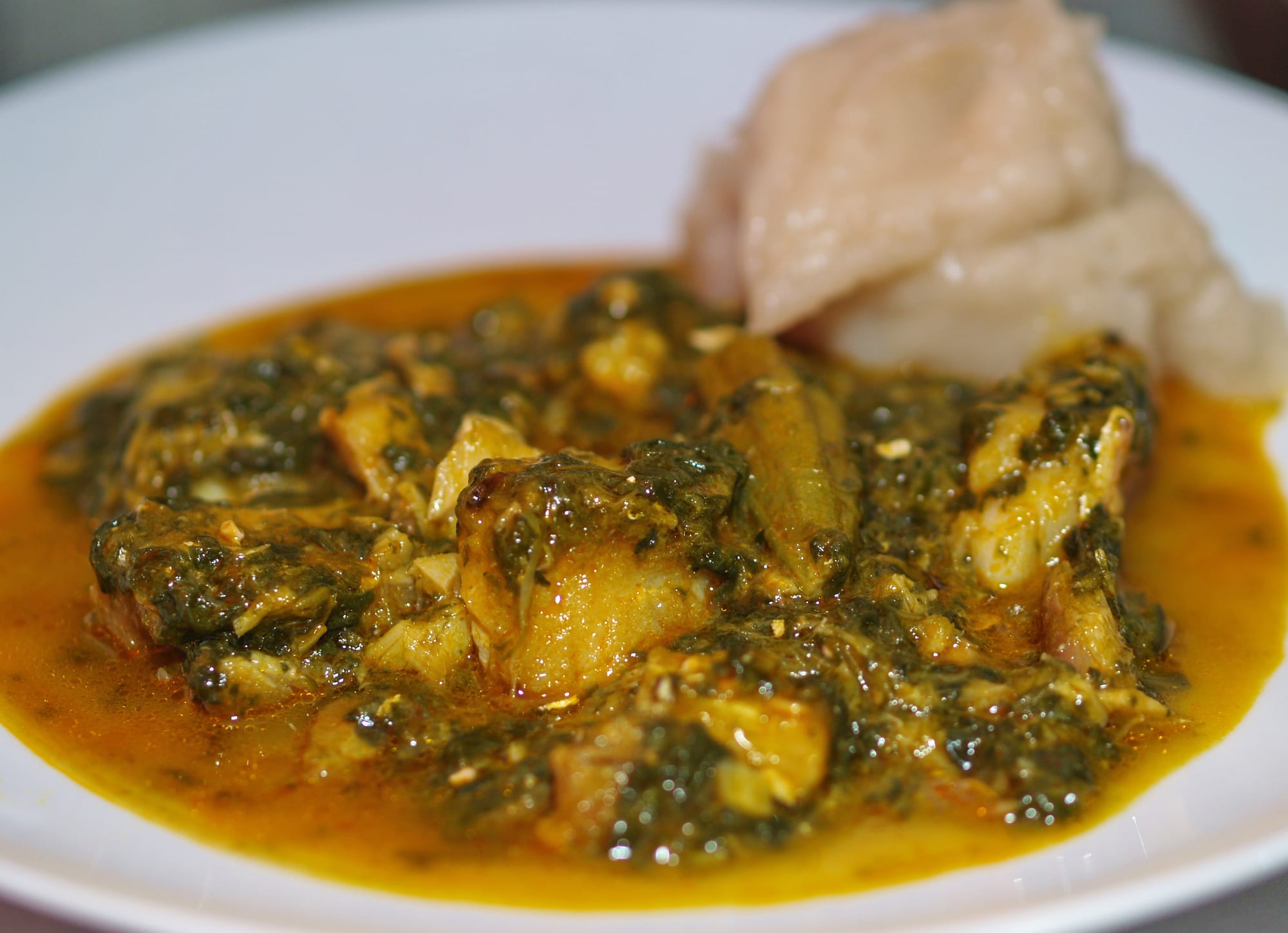 A square plate of fish and chicken pieces in a dark, oil-glossed green sauce, with white cassava starch, sliced plantain and okra pods