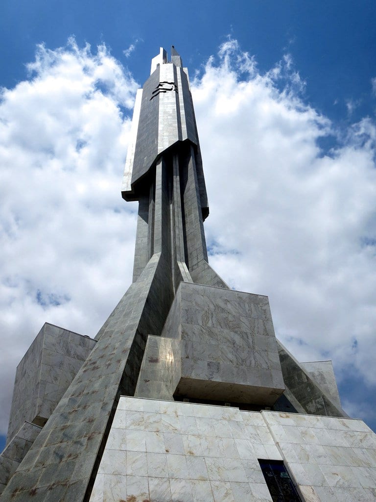 A tall pointed grey concrete tower shaped like a rocket, rising above low buildings