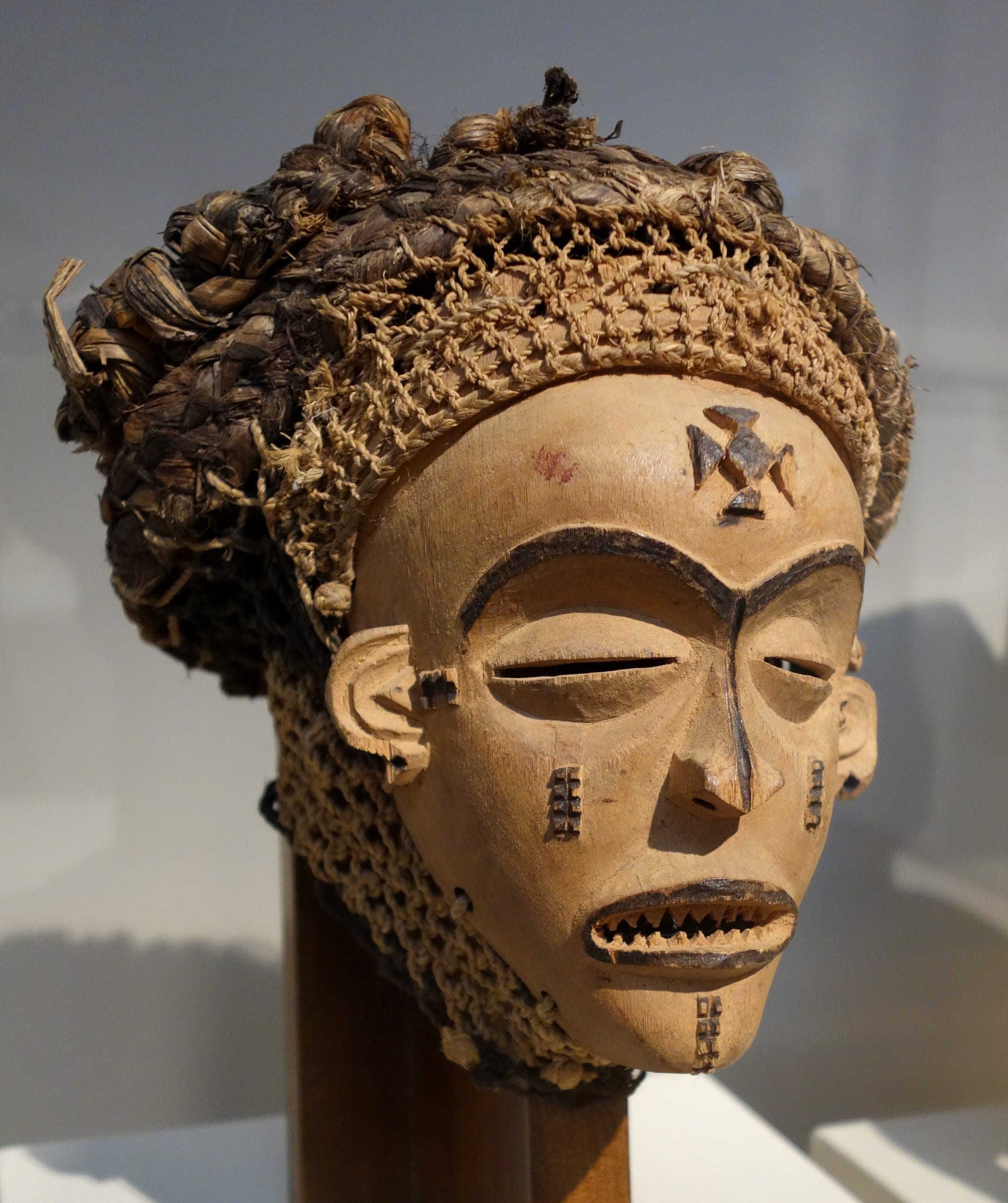 A carved pale wooden mask of a woman's face with narrow eyes, a cross-shaped mark on the forehead and a headdress of braided fibre and knotted twine
