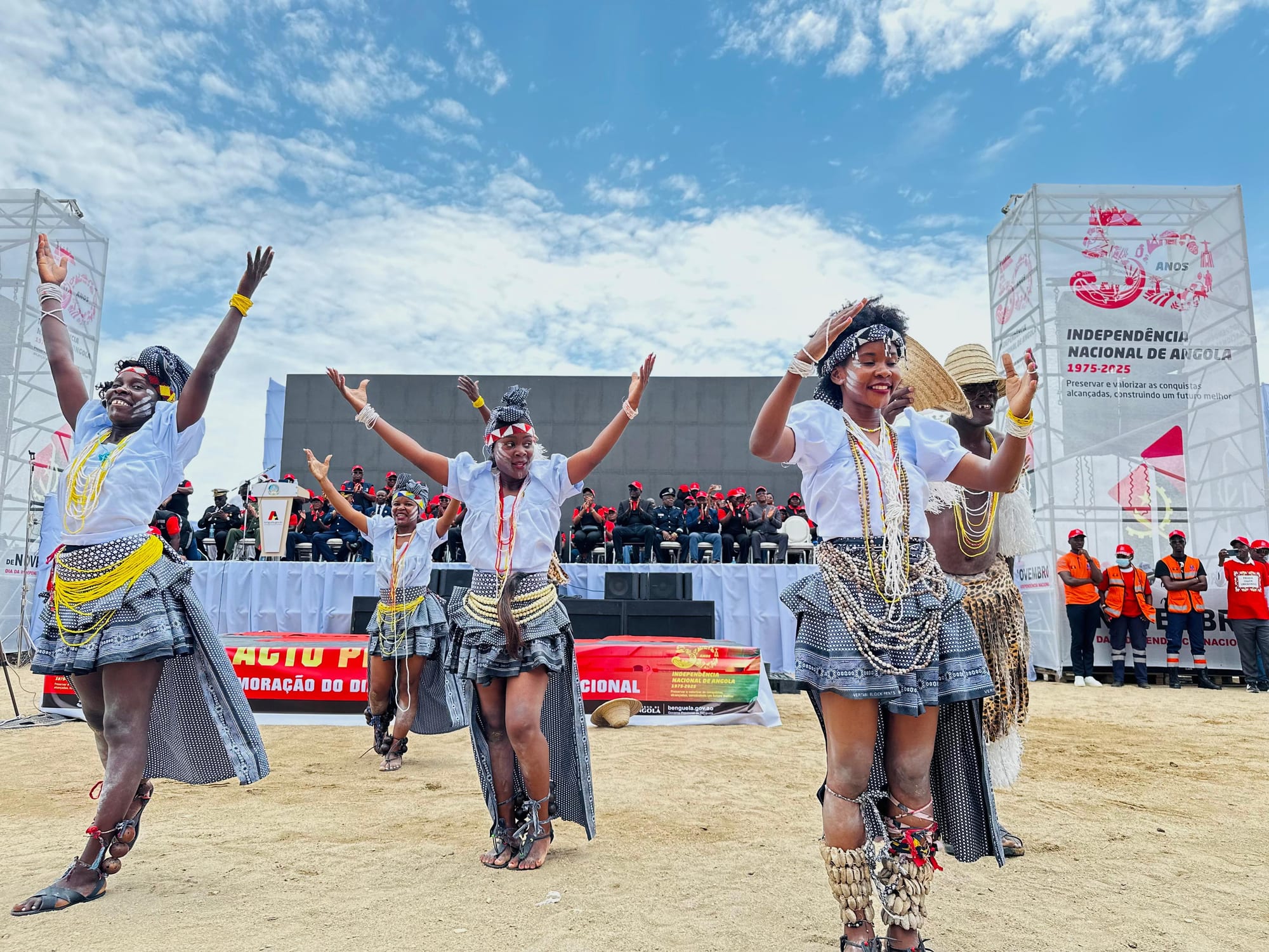 Four women dancers in white blouses, patterned skirts and bead necklaces dance with arms raised on open ground in front of a stage and a banner marking fifty years of Angolan independence