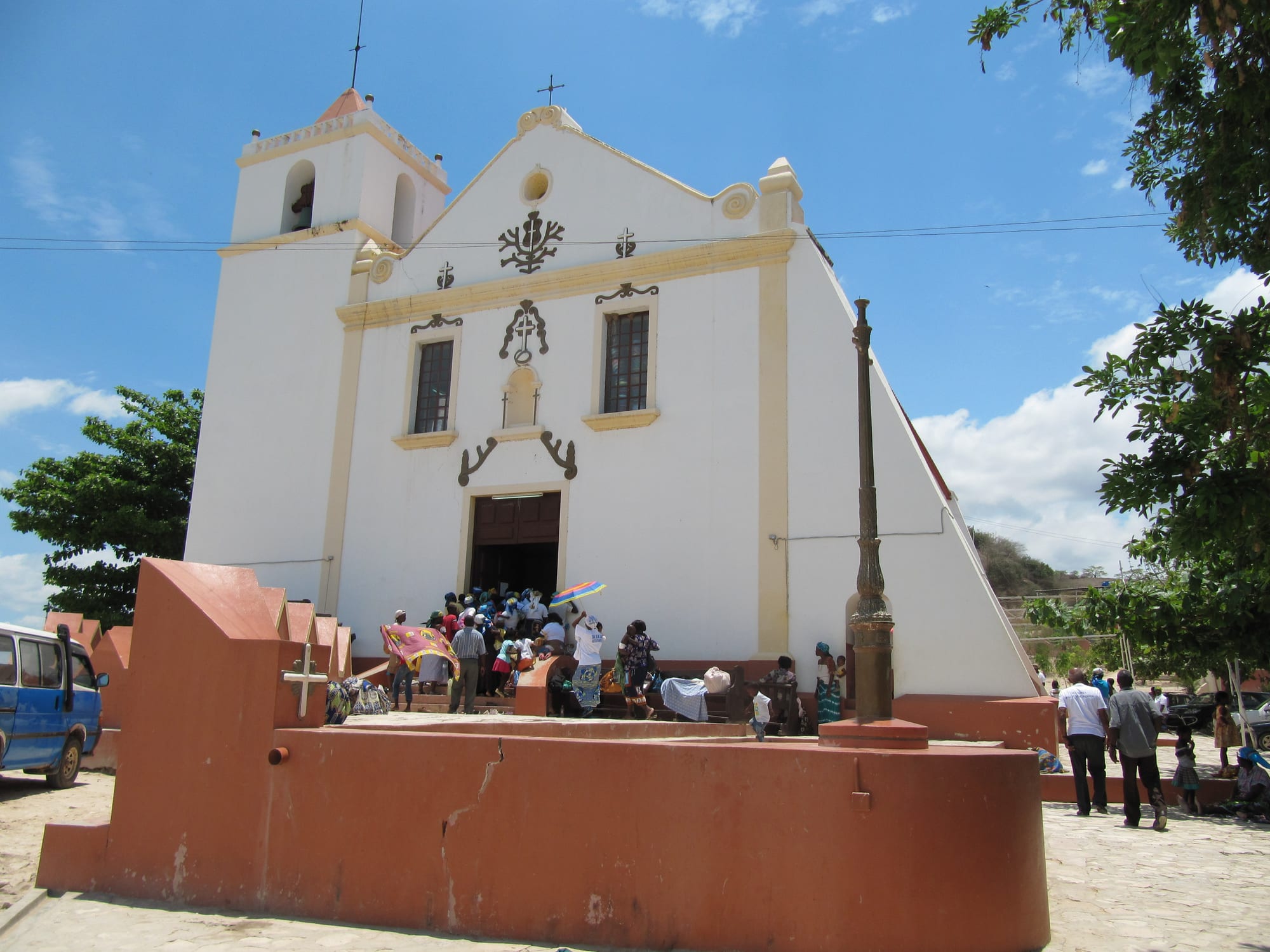 A white church with a bell tower and a stepped gable, with a crowd of visitors gathered at its open door and an orange-red wall in the foreground