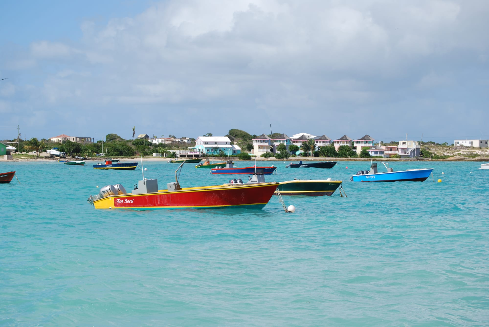 Colourful fishing and dive boats anchored in turquoise water off Island Harbour, Anguilla, with pastel village houses along the shore behind
