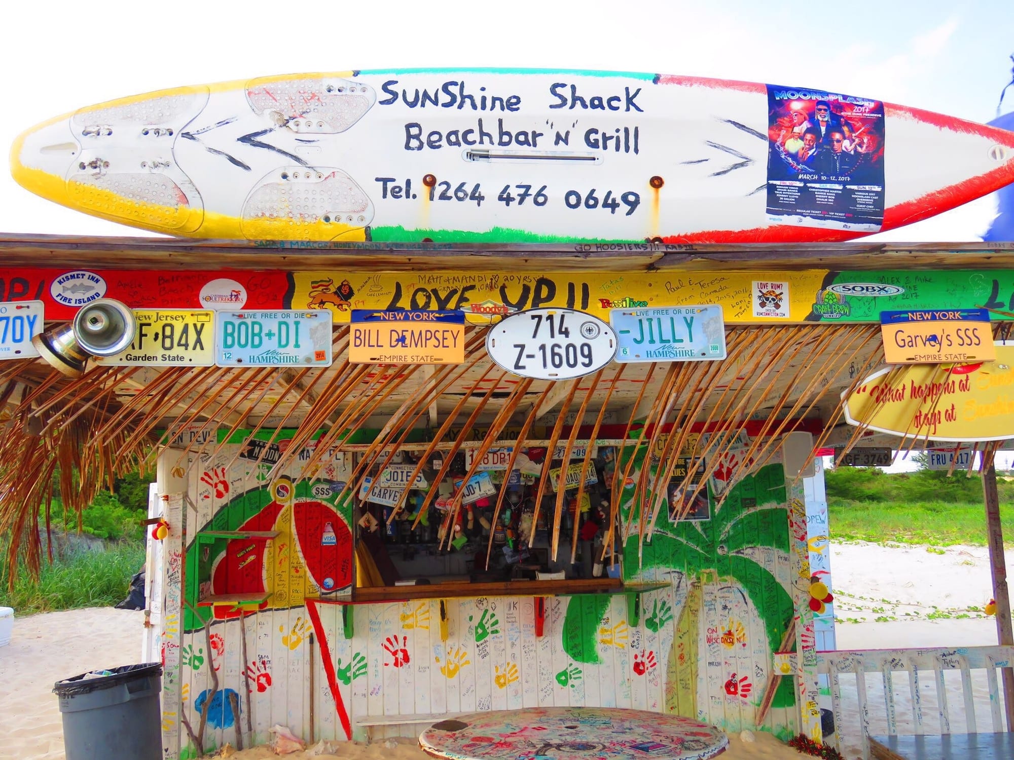 A hand-painted beach bar called Sunshine Shack, decorated with old car licence plates and a surfboard sign, on a sandy beach in Anguilla
