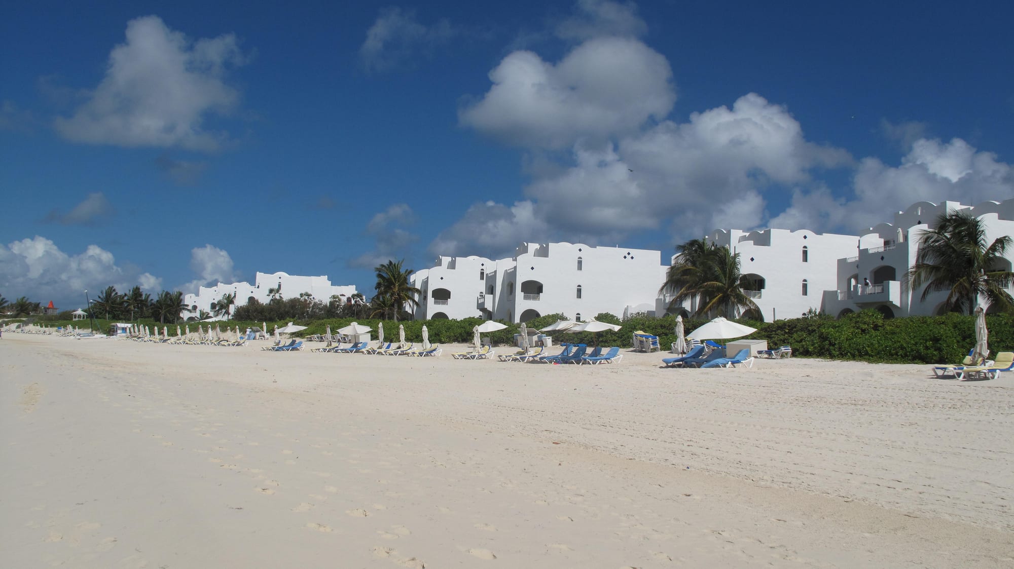 A wide sandy bay with gentle surf, low resort buildings set back among greenery