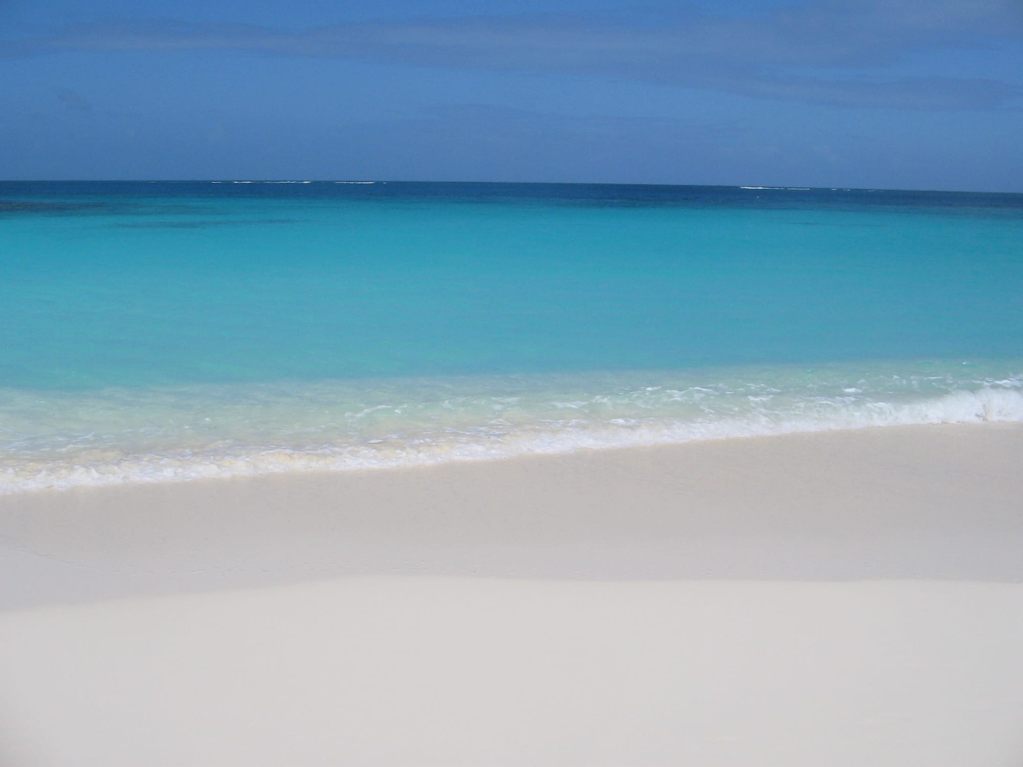 Pale turquoise shallows over white sand fading to deep blue, low green shore