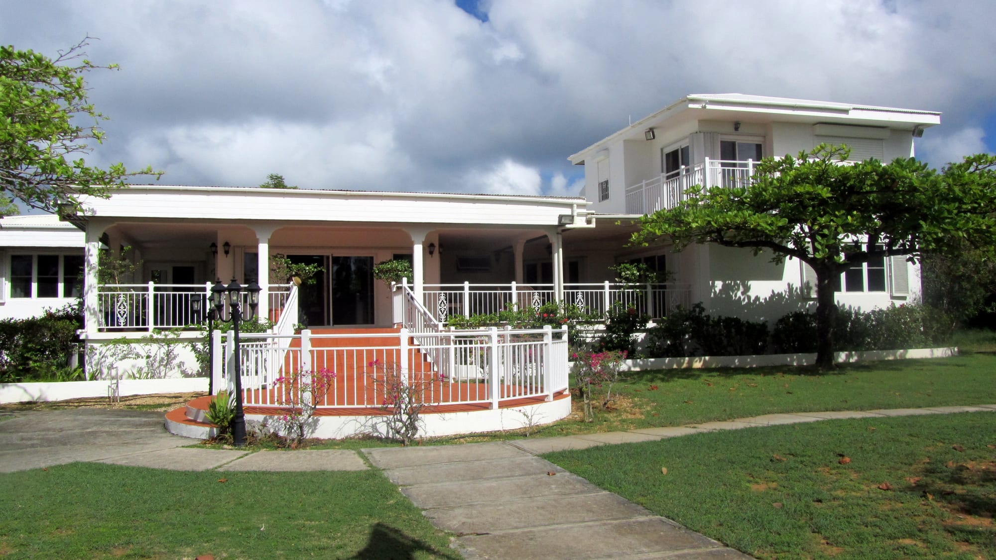 A low white house with a wide columned veranda, white railings and a red-tiled front terrace, set in lawns under a cloudy sky