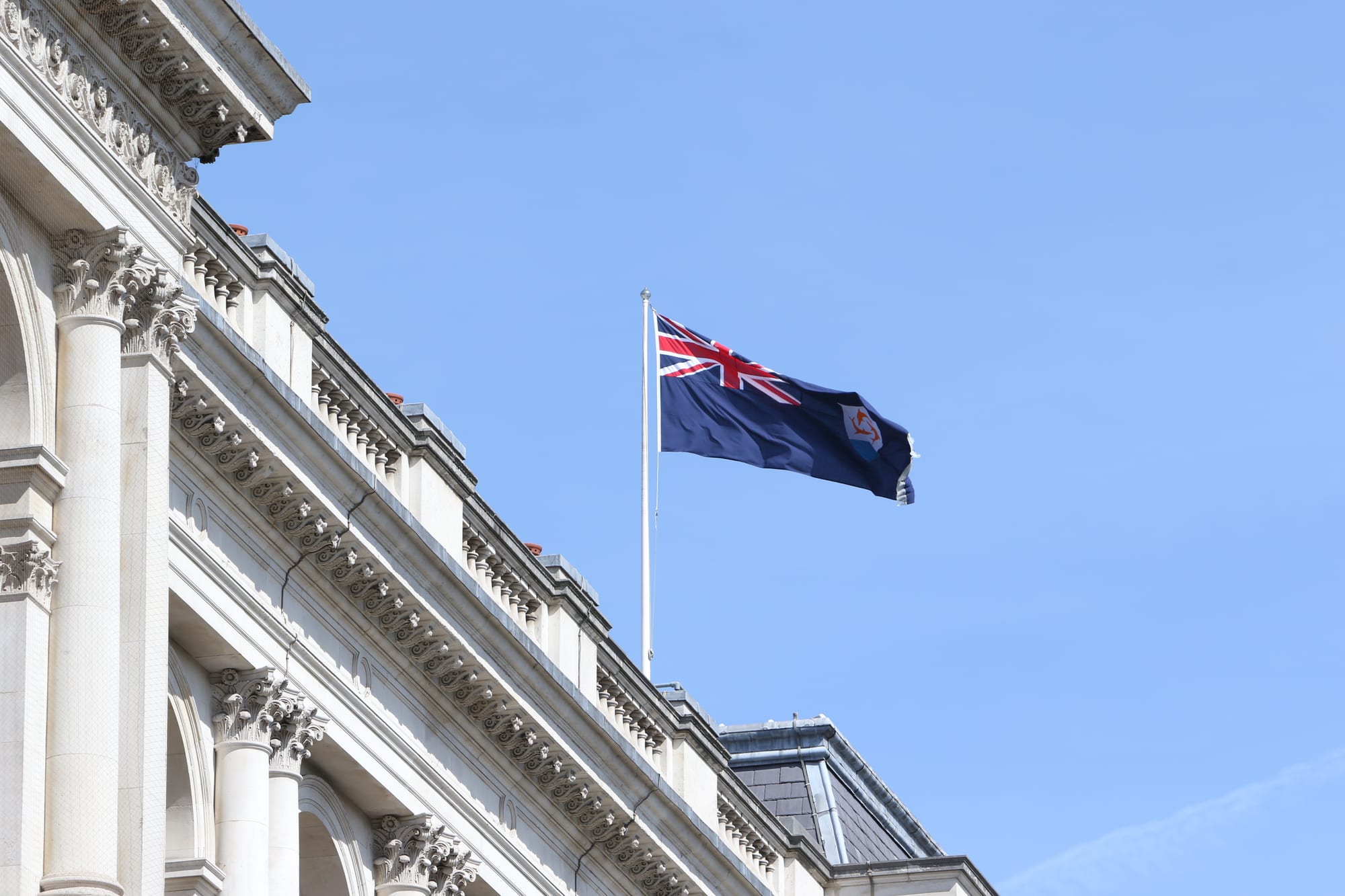 The flag of Anguilla, a dark blue ensign with the Union Flag in the corner and a white shield bearing three orange dolphins, flying from a pole on the roof of a pale stone building against a blue sky