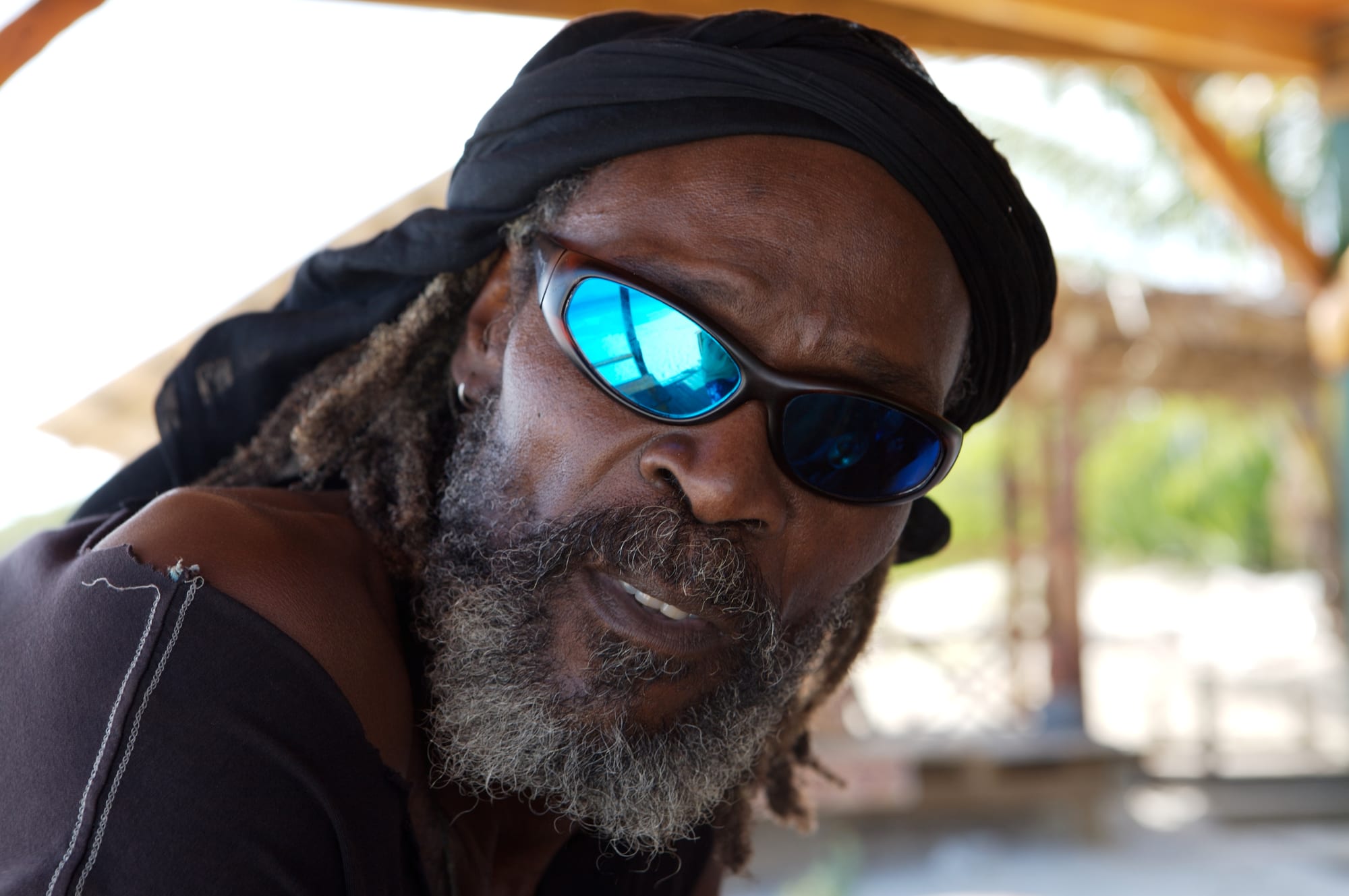 A close portrait of a bearded man with greying dreadlocks, a black head wrap and blue mirrored sunglasses, sitting under a wooden roof by a sandy beach