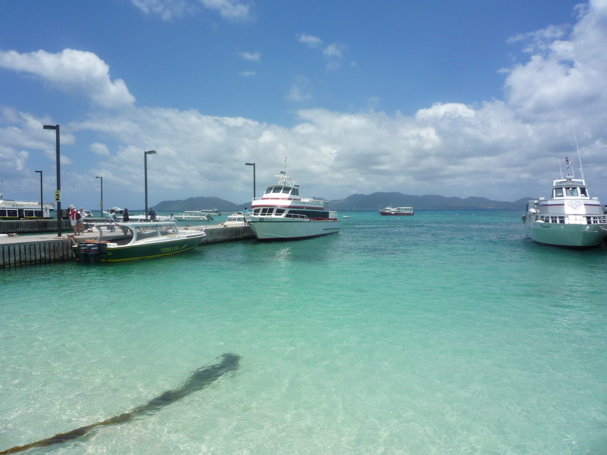 White passenger ferries and a small green boat moored beside a long concrete pier over clear turquoise water, with distant hills on the horizon under a blue sky with white clouds