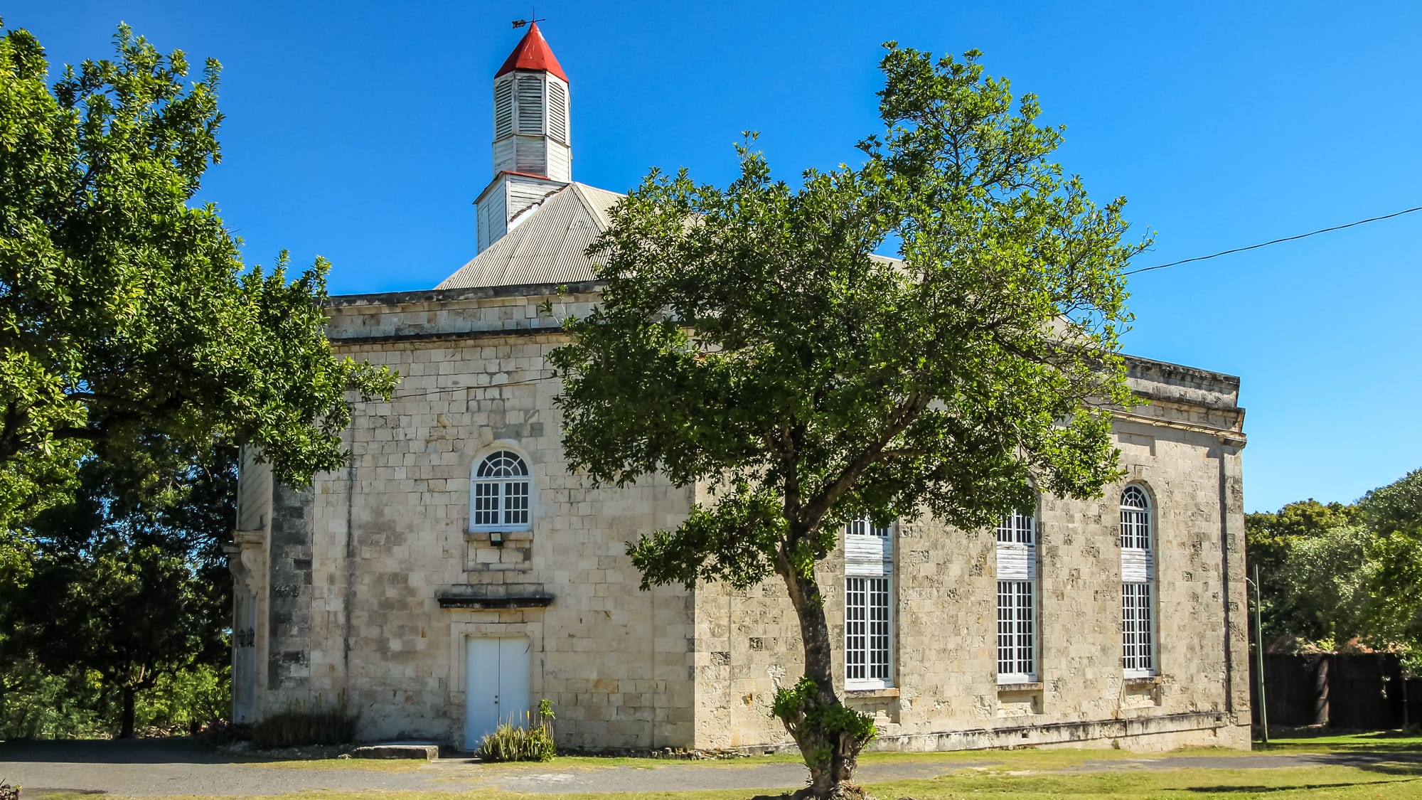 An octagonal stone church with tall arched windows and a small red-capped white cupola on its roof, with a leafy tree in front under a clear blue sky