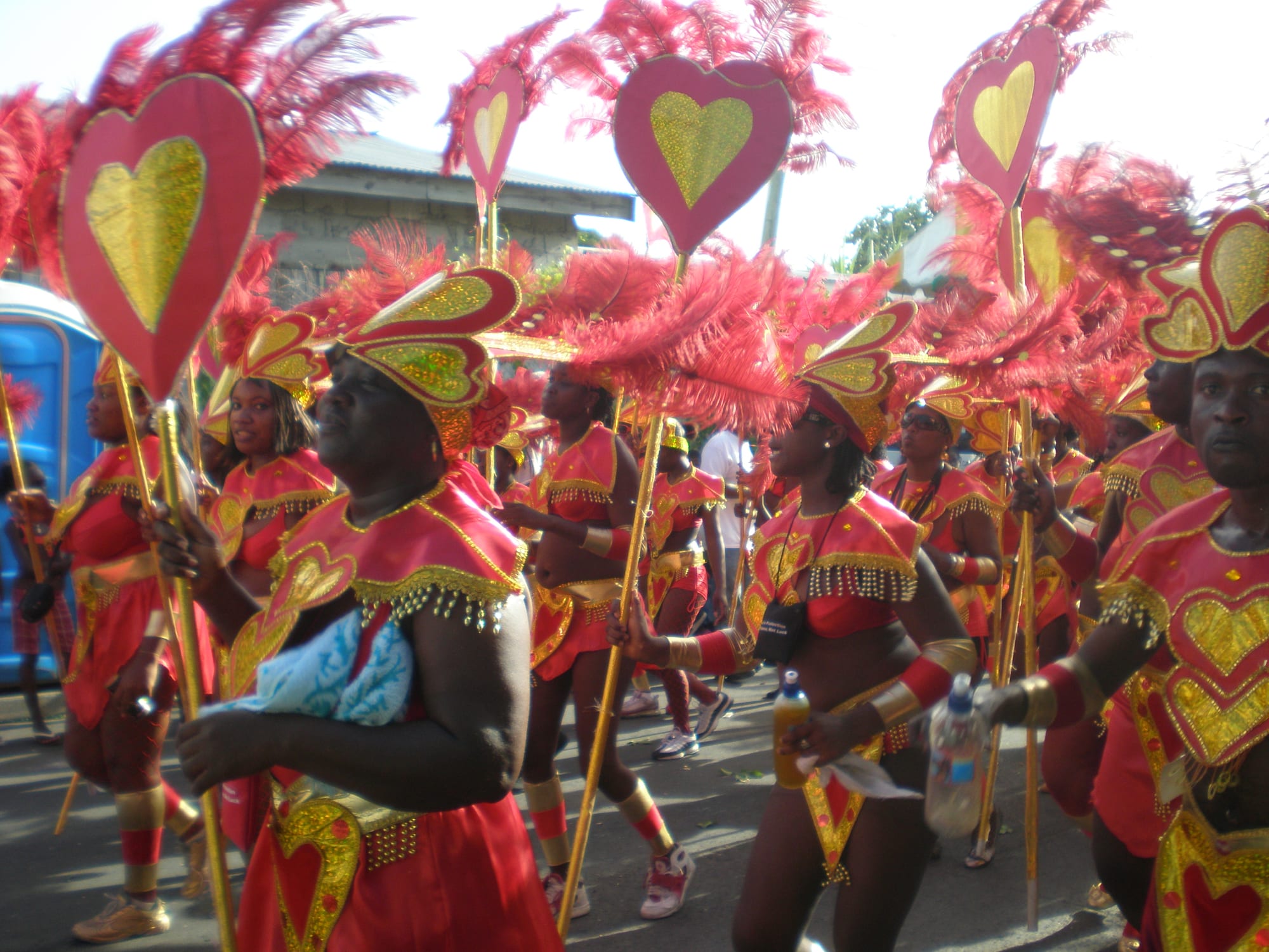 A troupe of carnival revellers in red and gold costumes with feathered headdresses and tall heart-shaped standards dancing down a street