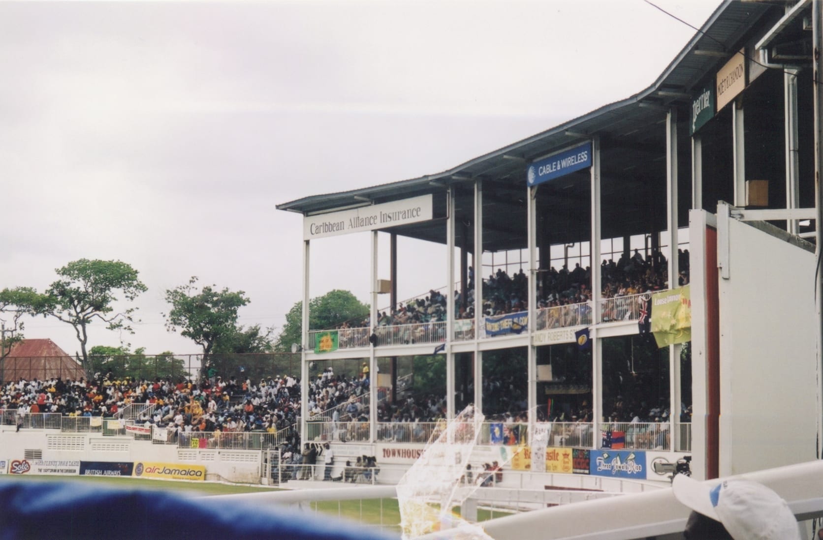 A crowded two-tier cricket stand with a corrugated roof and advertising boards, with more spectators packed along the lower terraces to the left under a grey sky