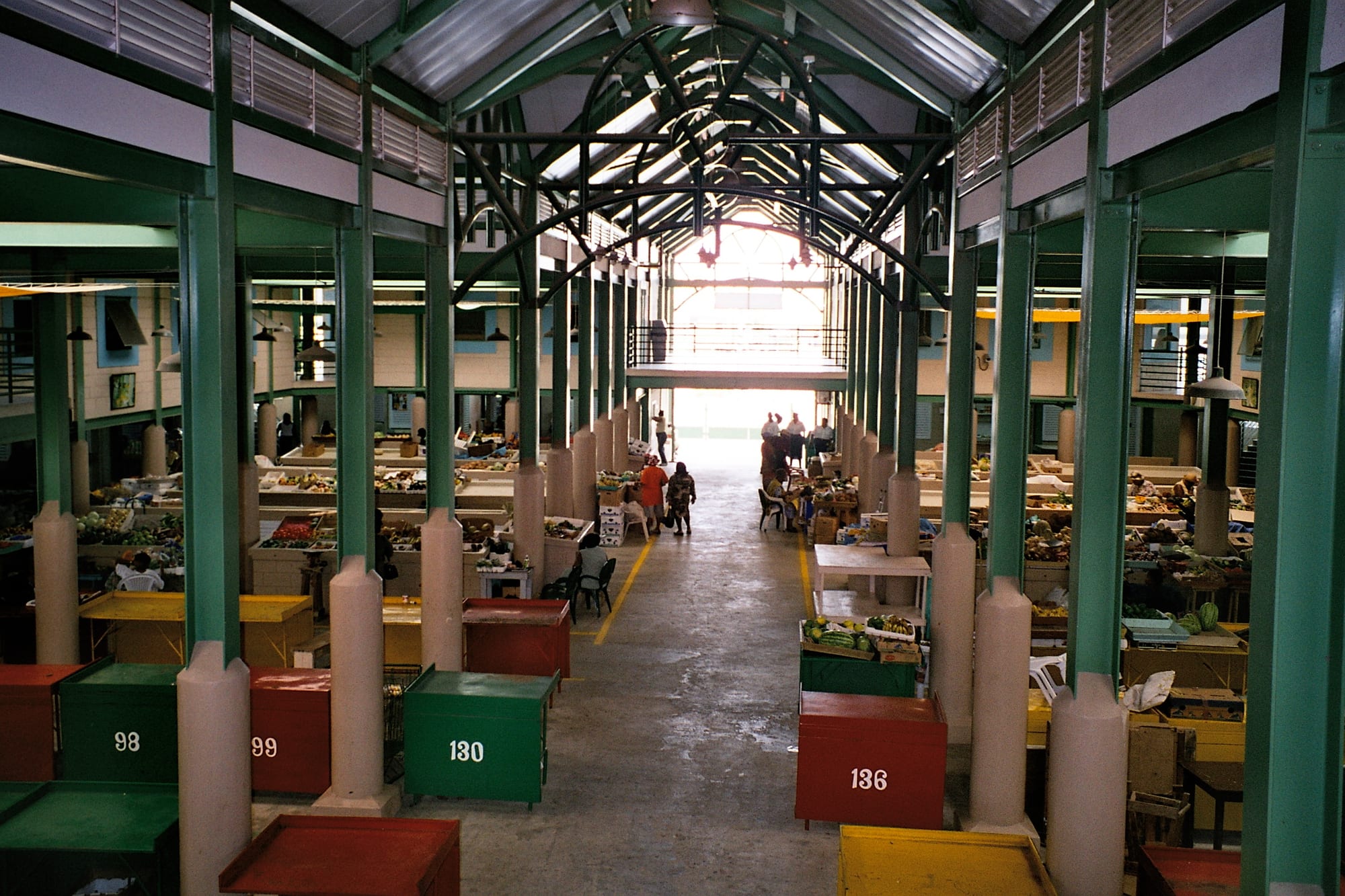 Rows of covered market stalls piled with fruit and vegetables under a green-painted steel roof, with a few shoppers and vendors along the aisle