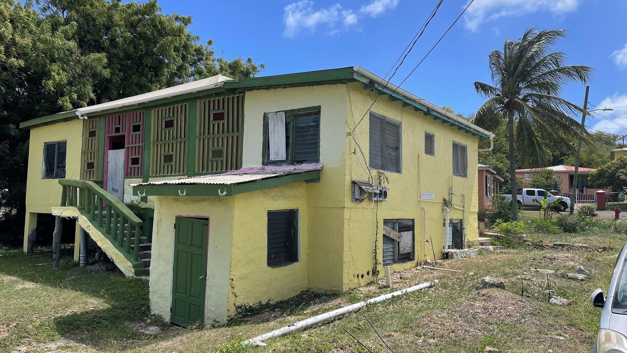 A yellow two-storey wooden house on low stilts with green shutters and an outside staircase, palm trees and a neighbouring house behind it