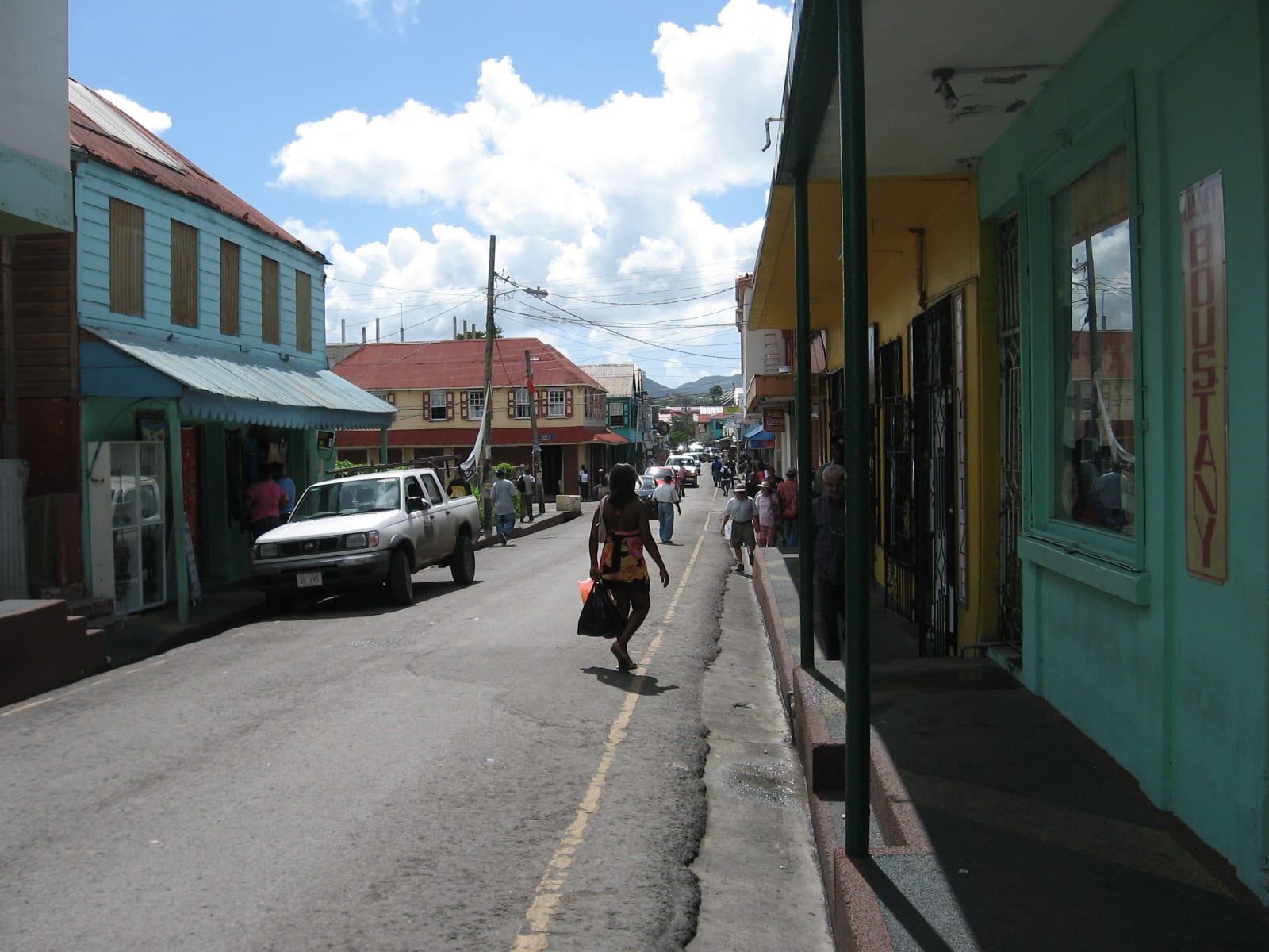 A woman walks down a street of colourfully painted wooden buildings in St John's, a pickup truck parked alongside and other pedestrians further along the pavement