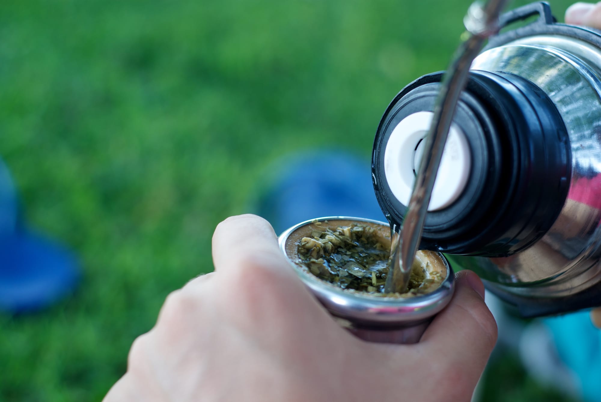 A hand holds a metal mate cup packed with yerba while a thermos pours hot water into it, with green grass in the background
