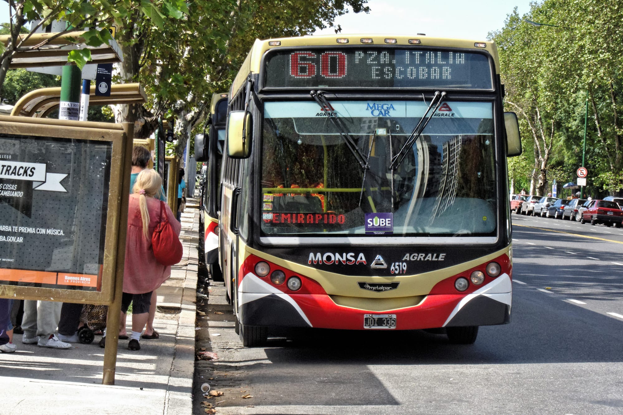 A red, cream and white city bus pulling up to a shaded bus stop where a woman waits with a shoulder bag