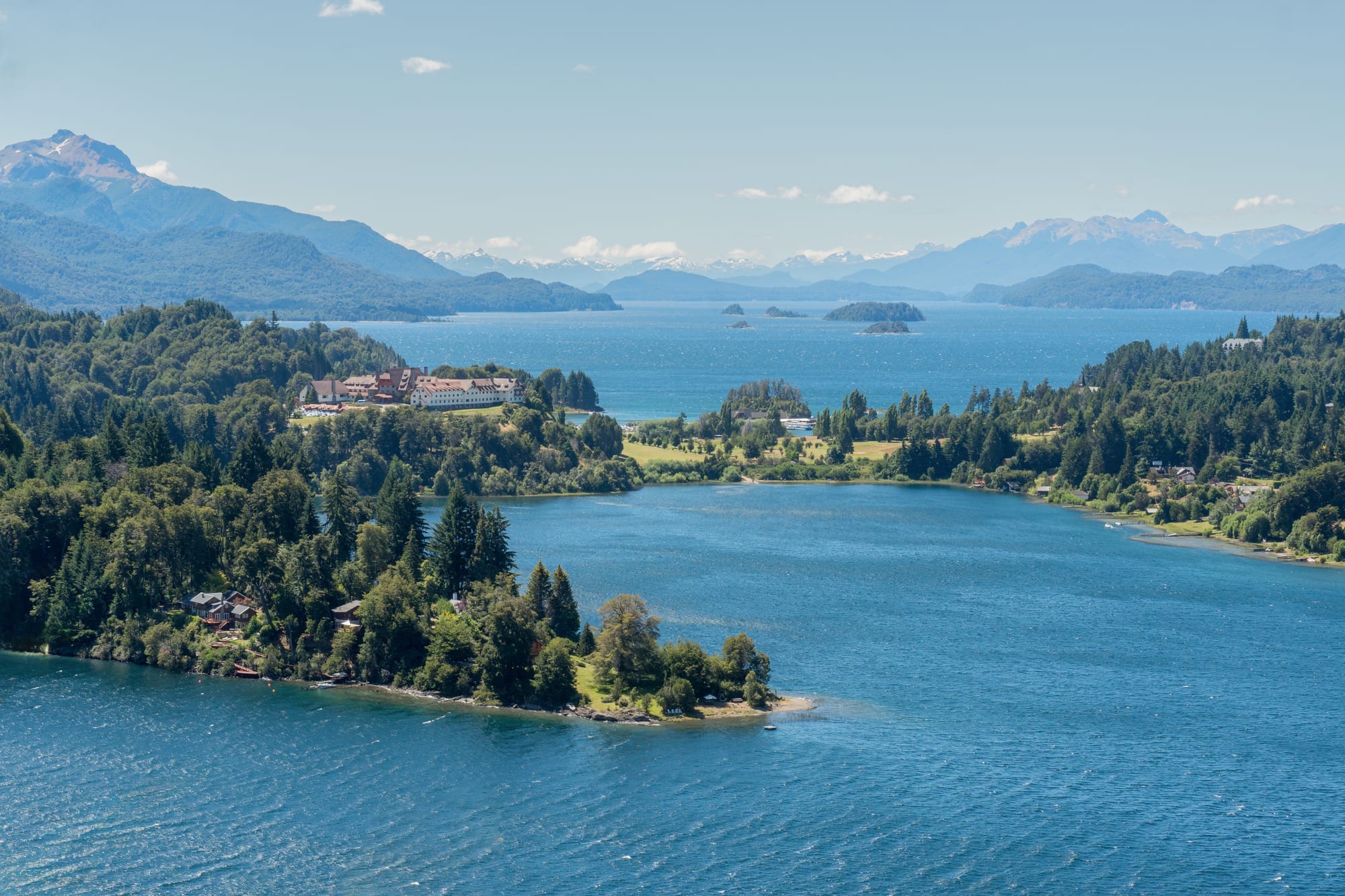 Forested peninsulas and islands in a deep-blue lake ringed by snow-capped mountains