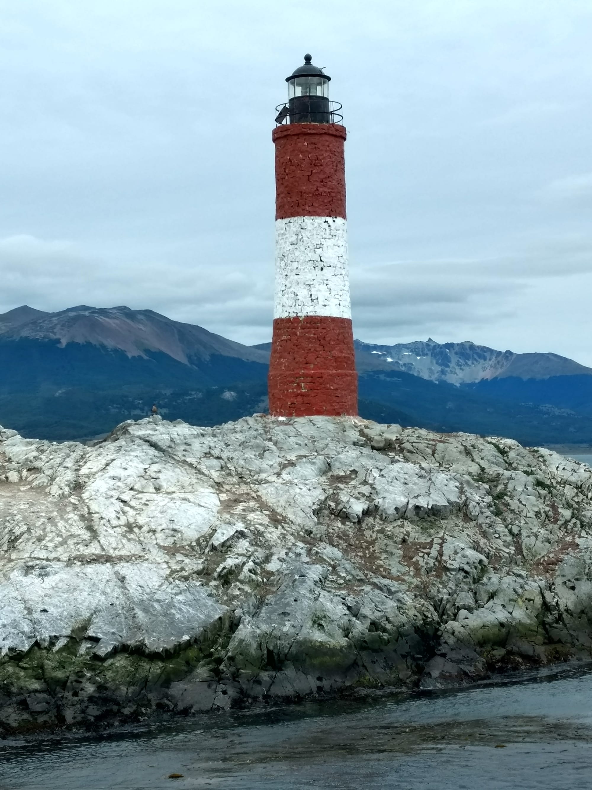 A small red-and-white lighthouse on a rocky islet in a channel below snow-dusted mountains