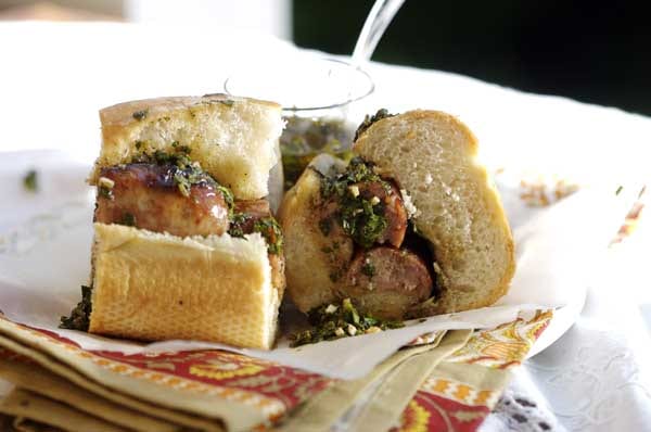 A grilled sausage split inside a crusty bread roll and topped with a green herb sauce, cut in half on a plate