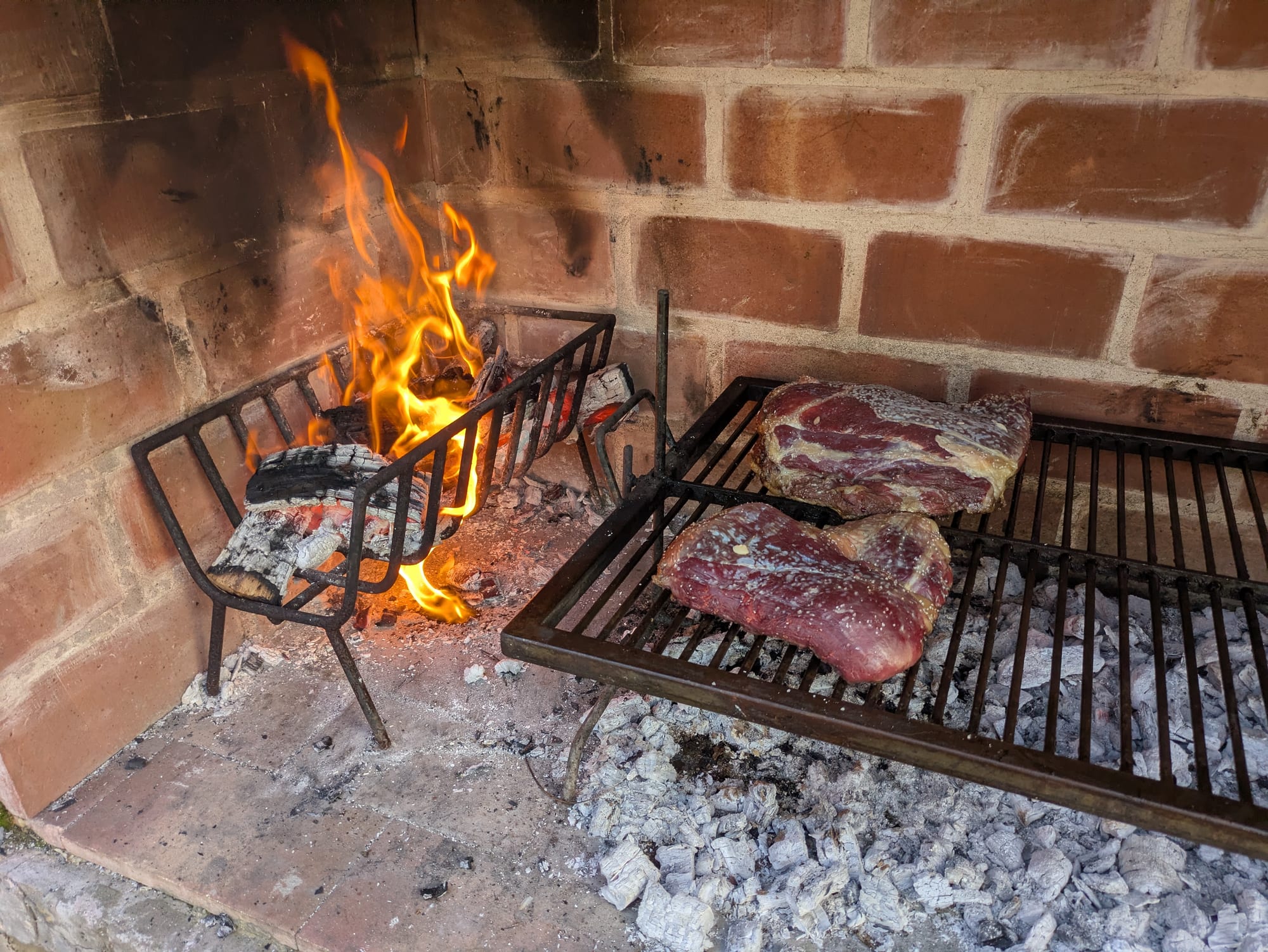 A brick barbecue with a wood fire burning on one side and two large cuts of beef laid on an iron grill beside the embers