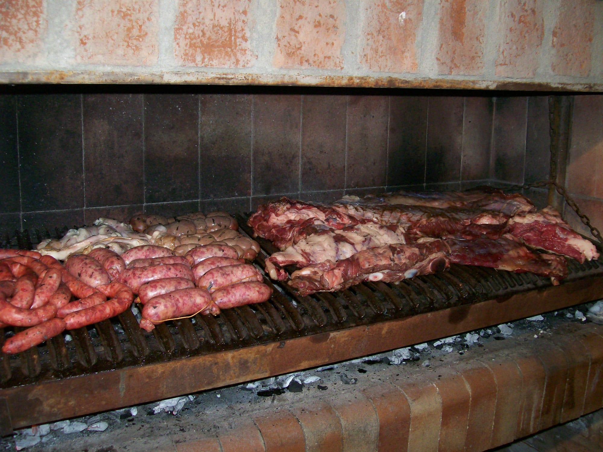 Sausages, offal and large cuts of beef ribs laid out on a long built-in brick grill over glowing coals