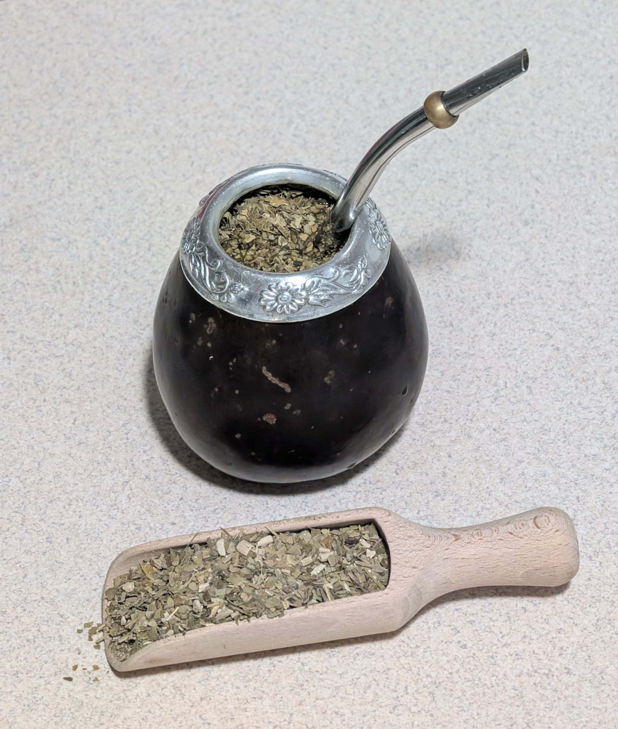 A dark gourd filled with loose green yerba mate leaves, fitted with a metal straw, beside a small wooden scoop of more yerba mate