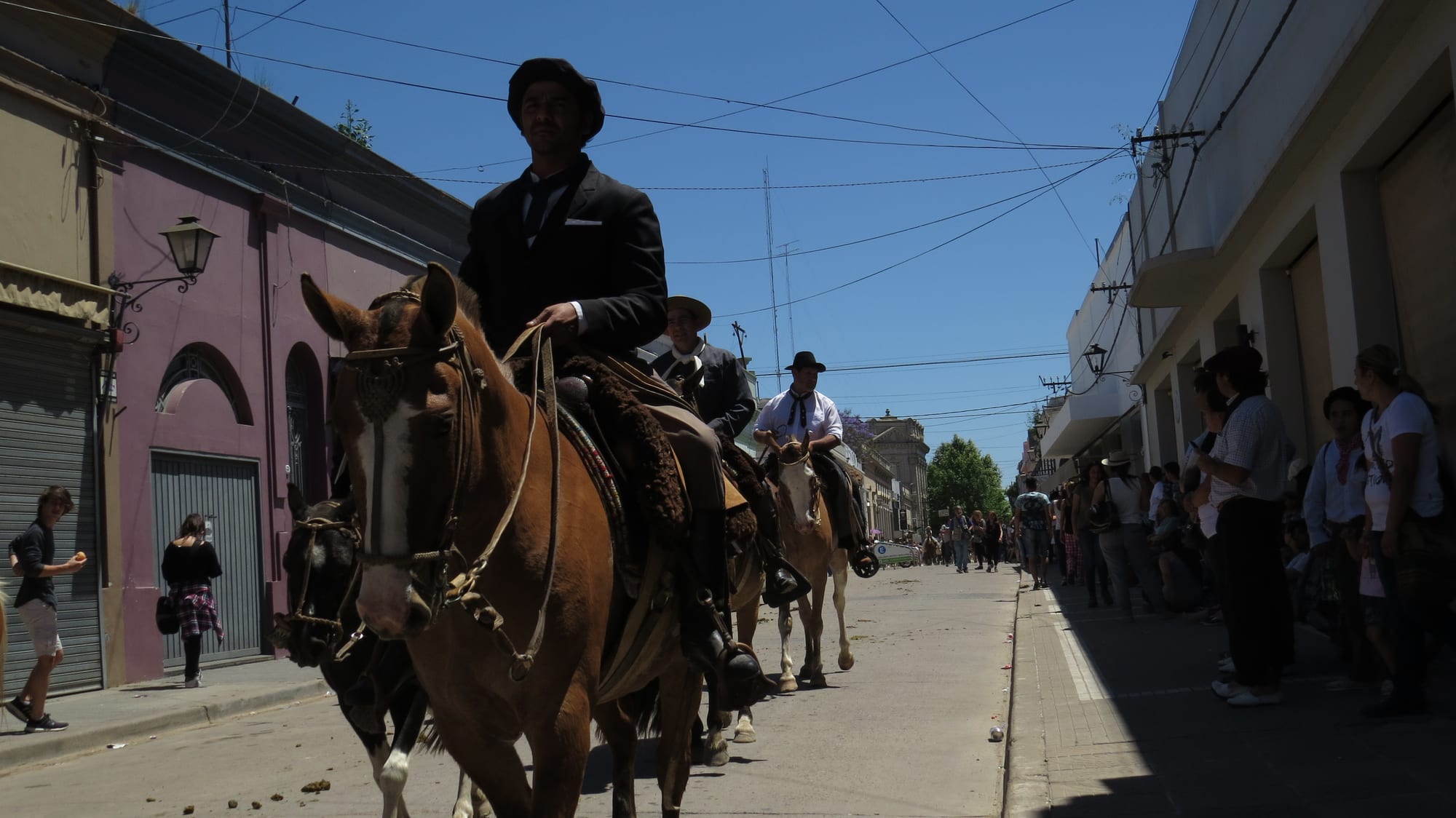 Riders in black berets, dark jackets and neckerchiefs ride horses with sheepskin saddles down a sunny street lined with low buildings and spectators