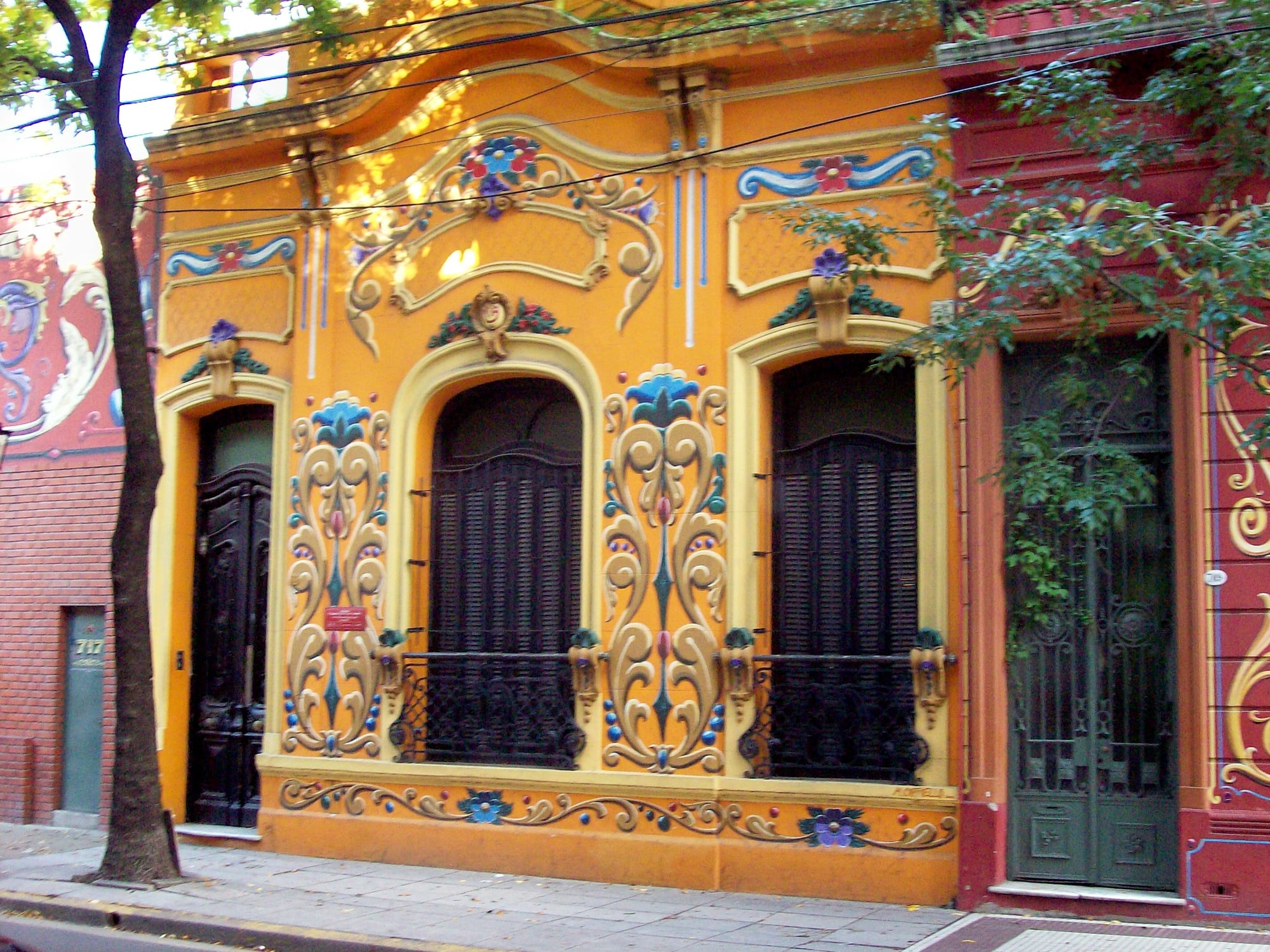 The orange facade of a single-storey house decorated with painted scrolls, flowers and curling ornaments in cream, blue, green and red around dark shuttered windows and doors