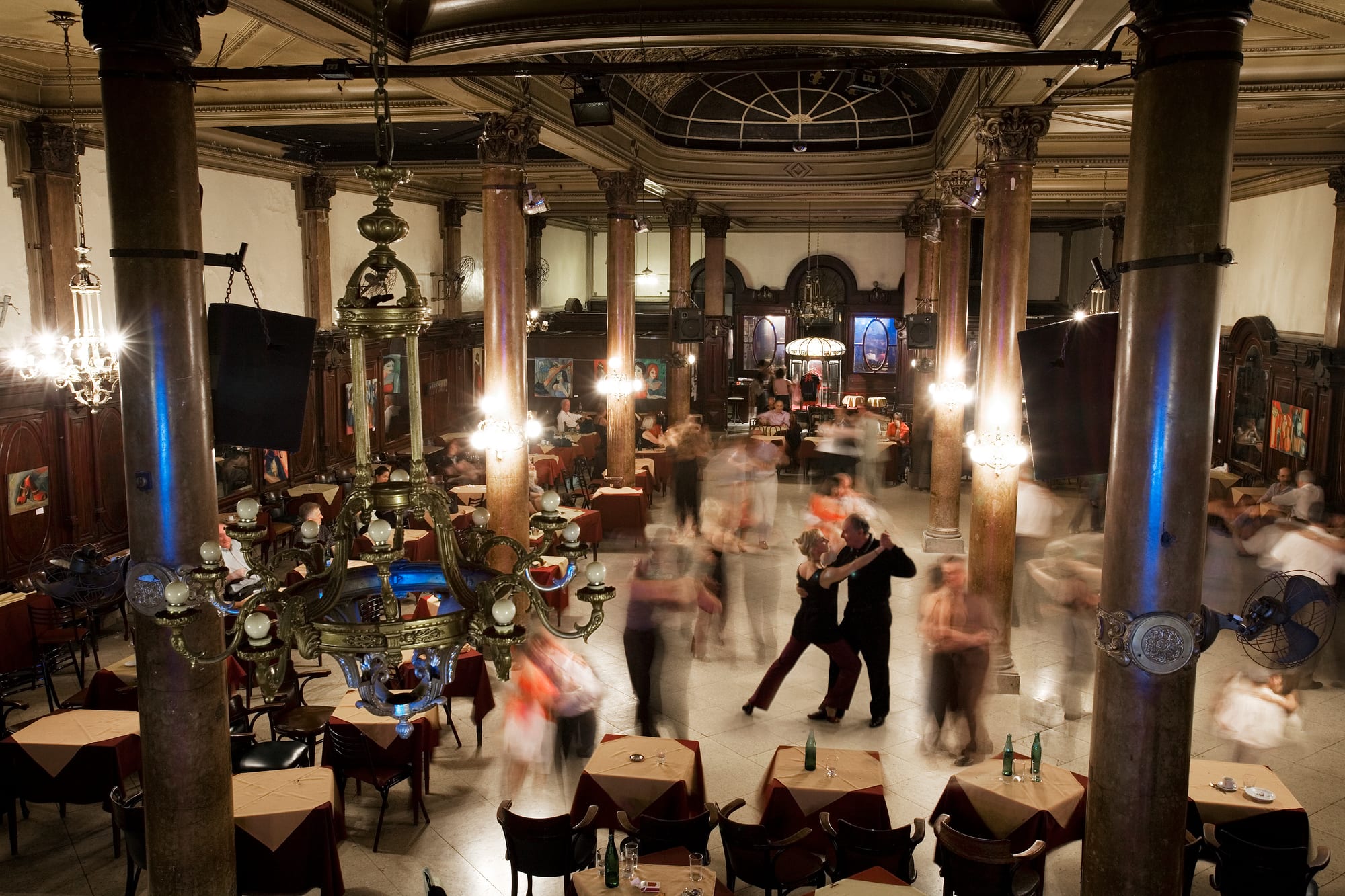 Couples dancing tango on a pale marble floor between tall marble columns and tables with red and white cloths, lit by ornate chandeliers in a grand old ballroom
