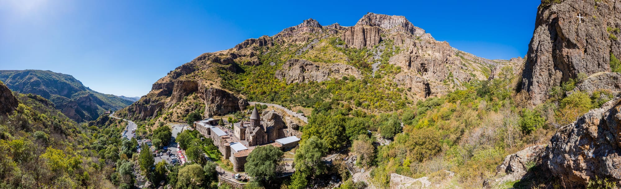 A medieval church complex of grey stone tucked against a tall cliff in a narrow valley
