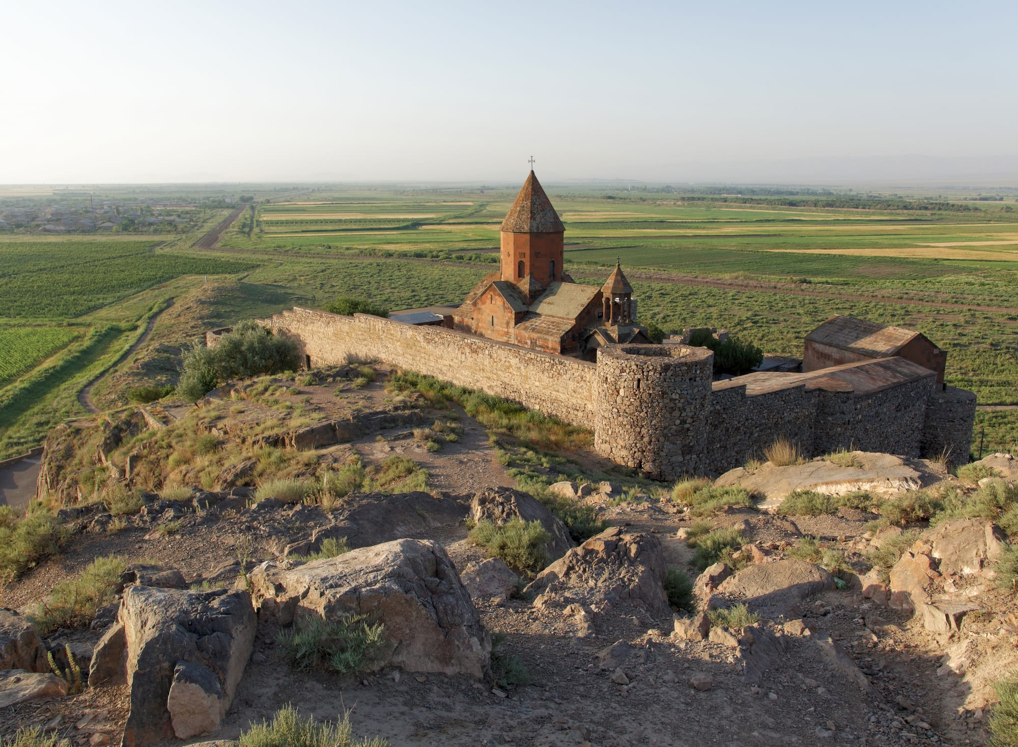 A small fortified monastery on a knoll on a flat plain, snowy Ararat filling the sky behind