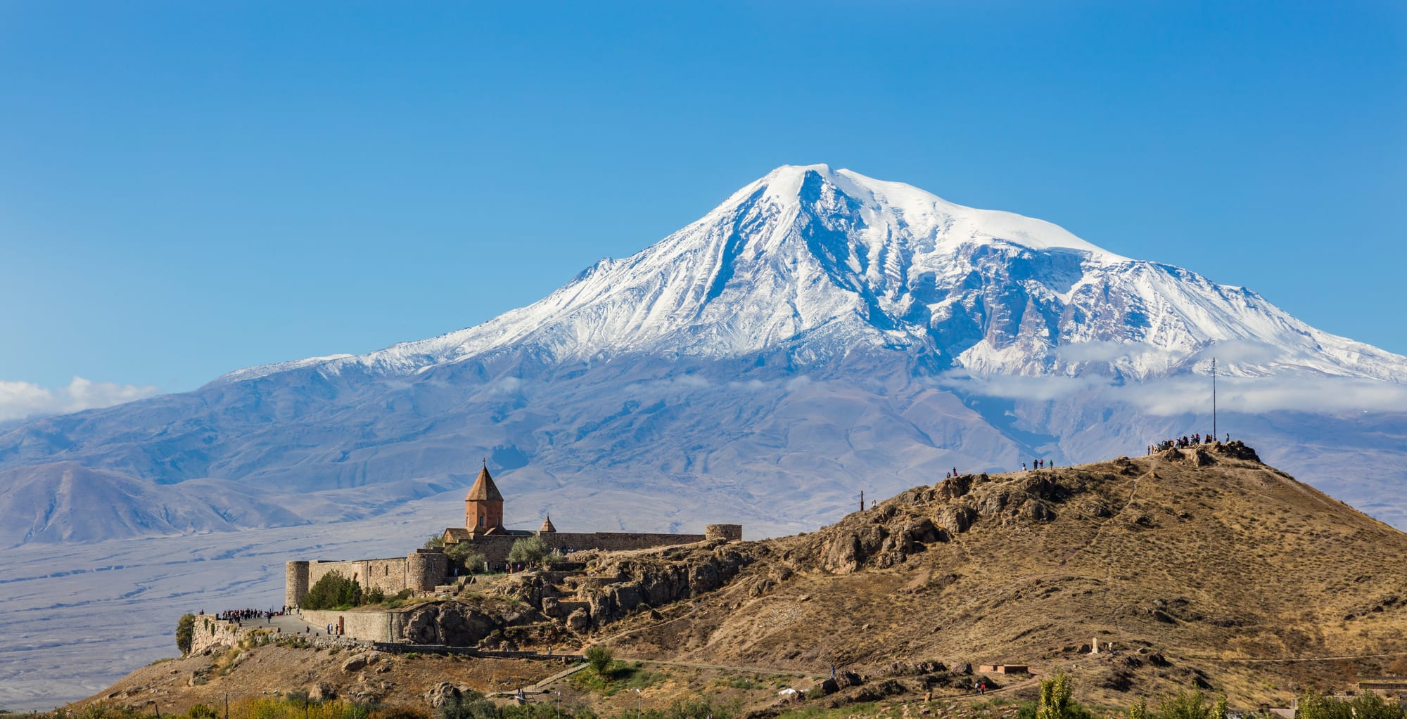 A walled monastery on a low hill with the huge snow-capped cone of Mount Ararat behind it