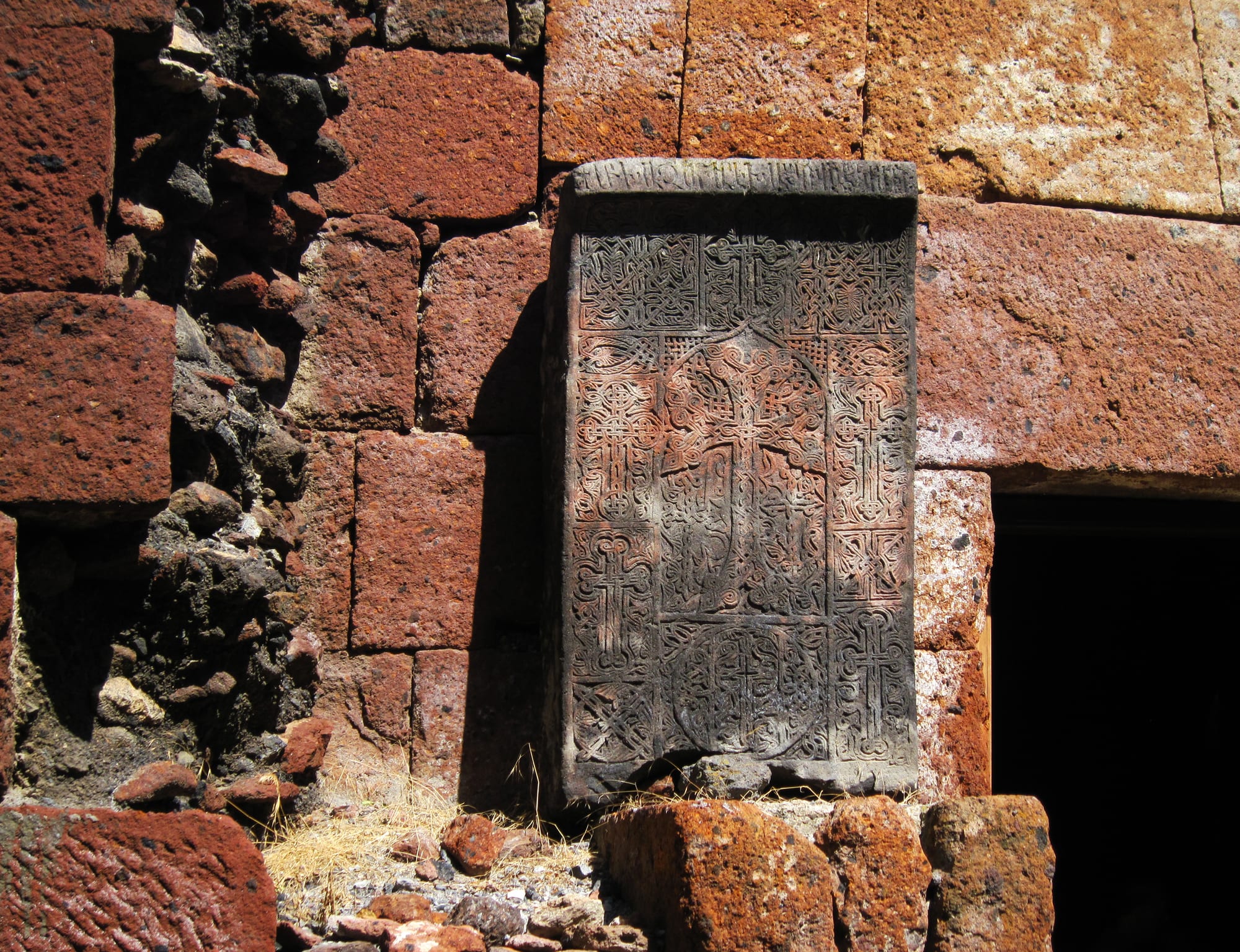 A grey stone slab carved all over with a cross and dense interlaced patterns, set into a wall of large red and dark volcanic stone blocks