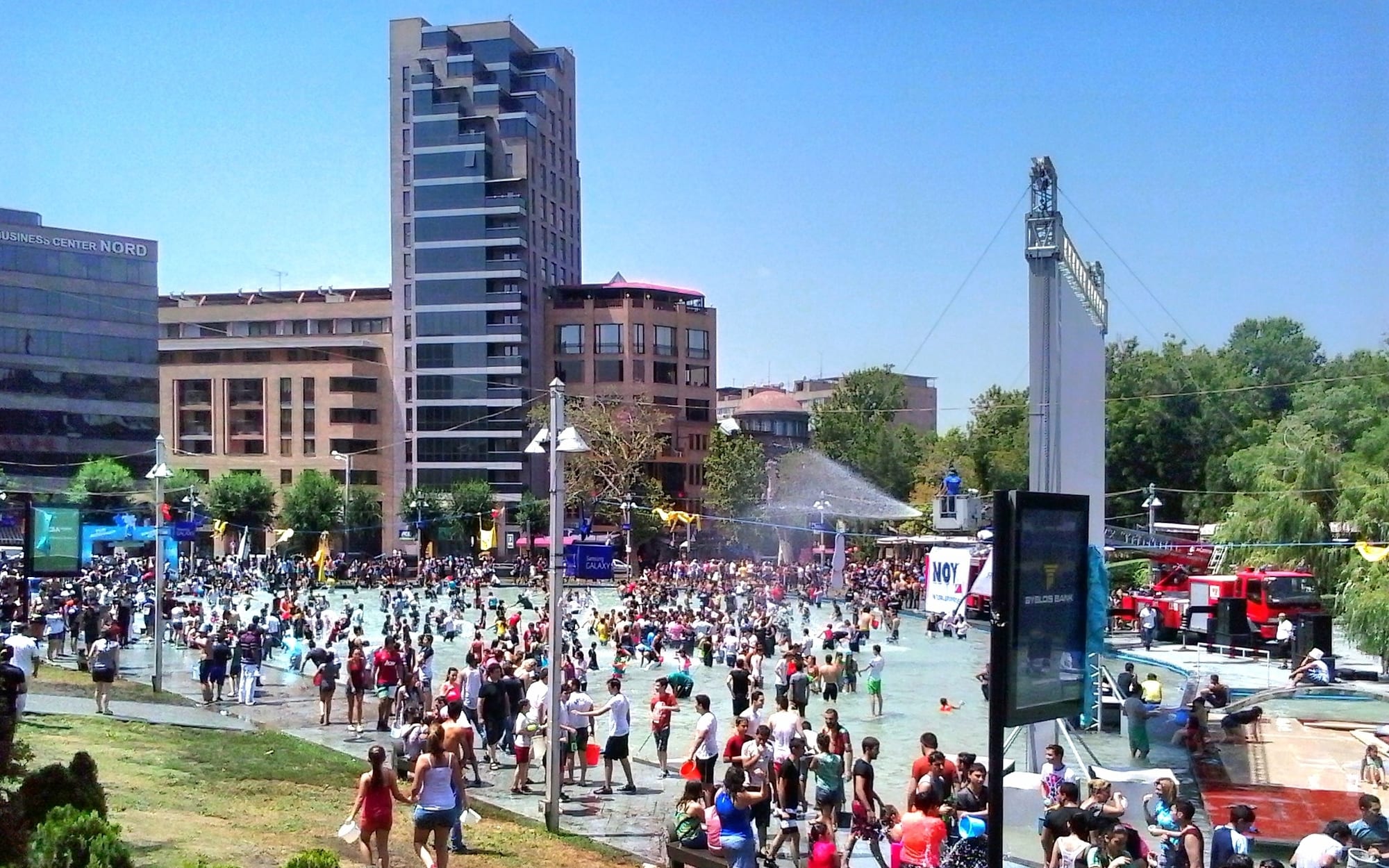 A large crowd of people in summer clothes wading and splashing in a wide shallow fountain square among city buildings, with a red fire engine on the right