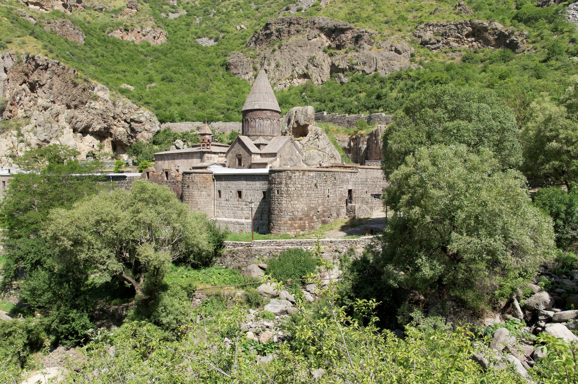 A medieval stone monastery with a conical-domed church and thick round-cornered walls, surrounded by green trees below rocky cliffs
