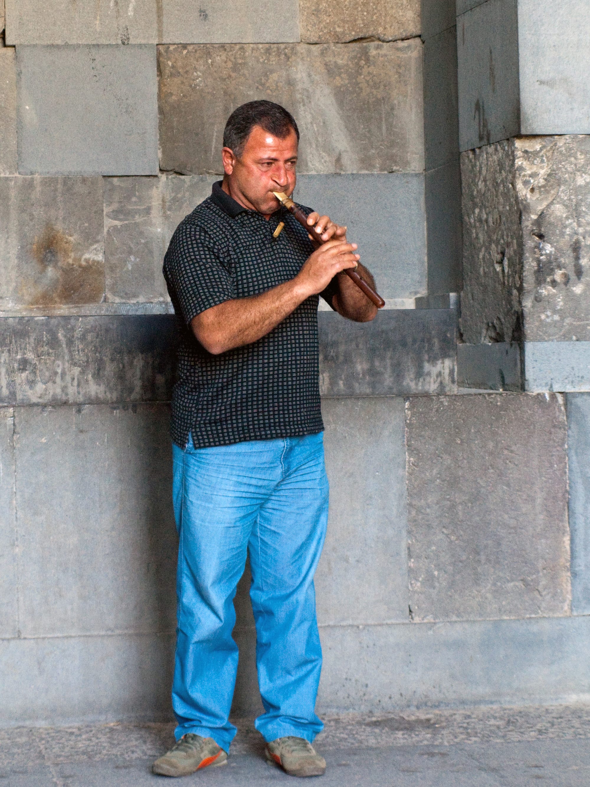 A man in a dark checked shirt and blue jeans plays a short dark wooden pipe while standing against a wall of large grey stone blocks
