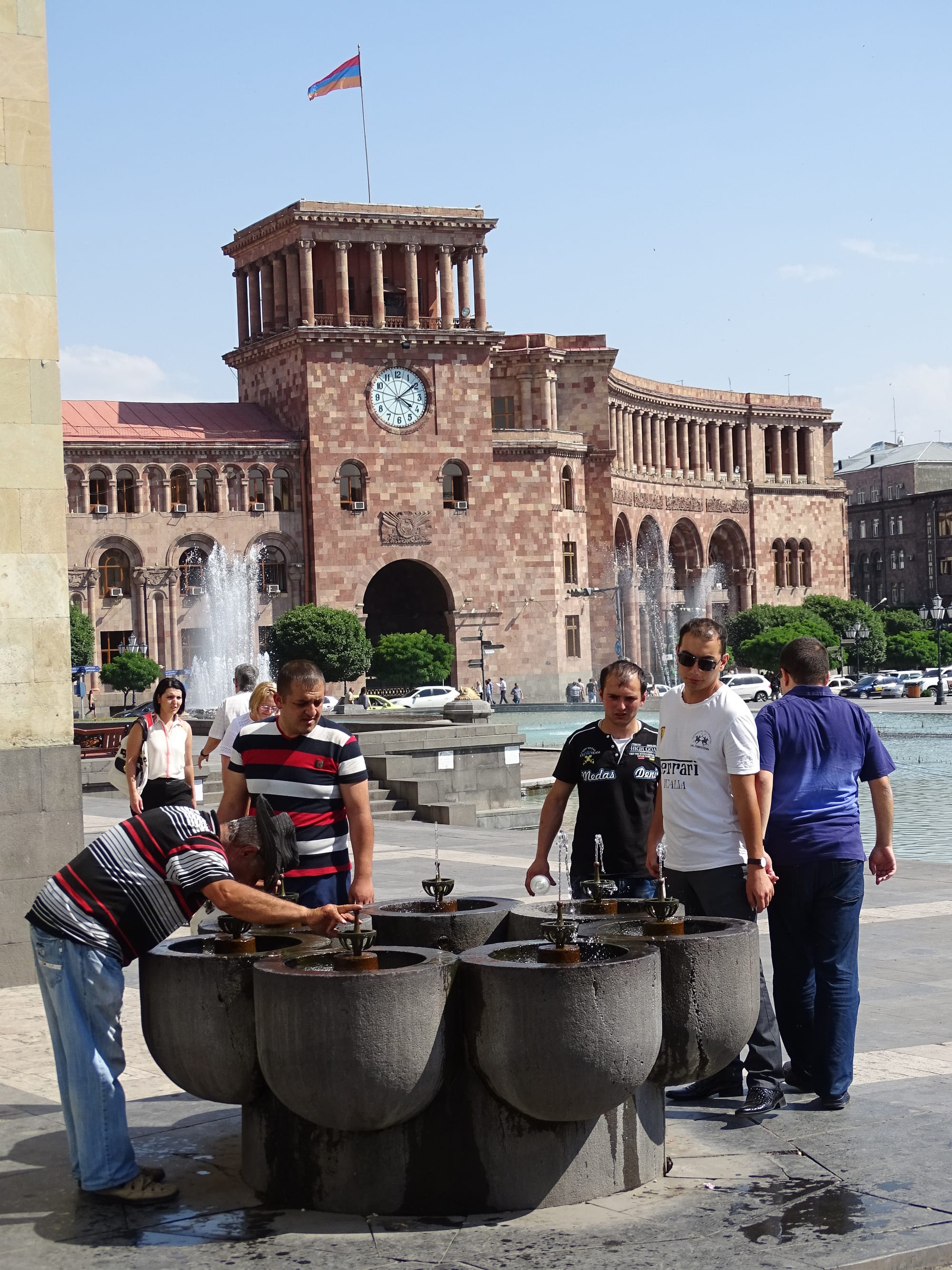 Several men stop to drink from a stone public drinking fountain in Yerevan's Republic Square, with the pink-toned Government House and its fountains behind them