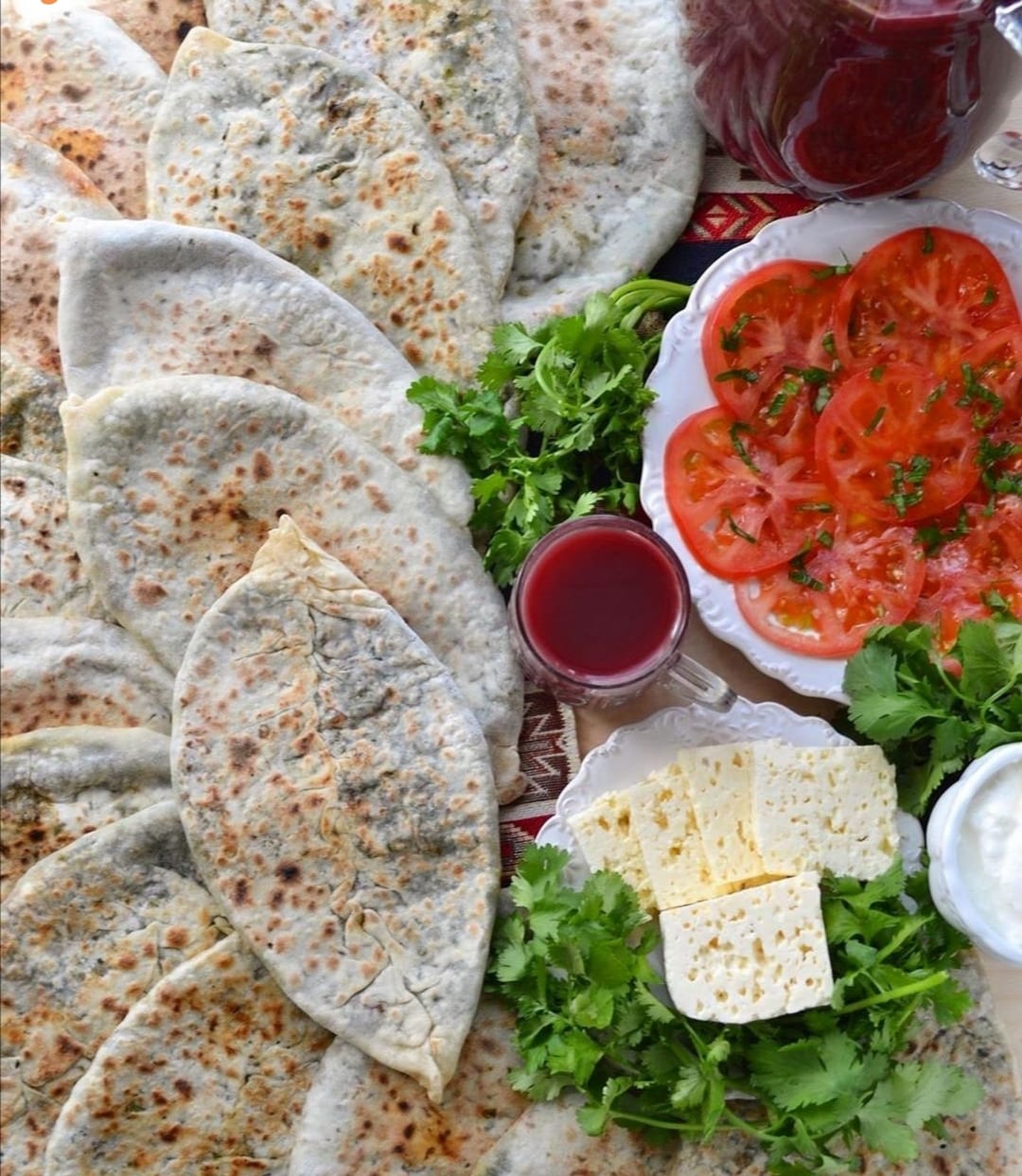 Folded flatbreads with griddle marks, filled with green herbs, served with cheese, tomato salad and a glass of dark red juice